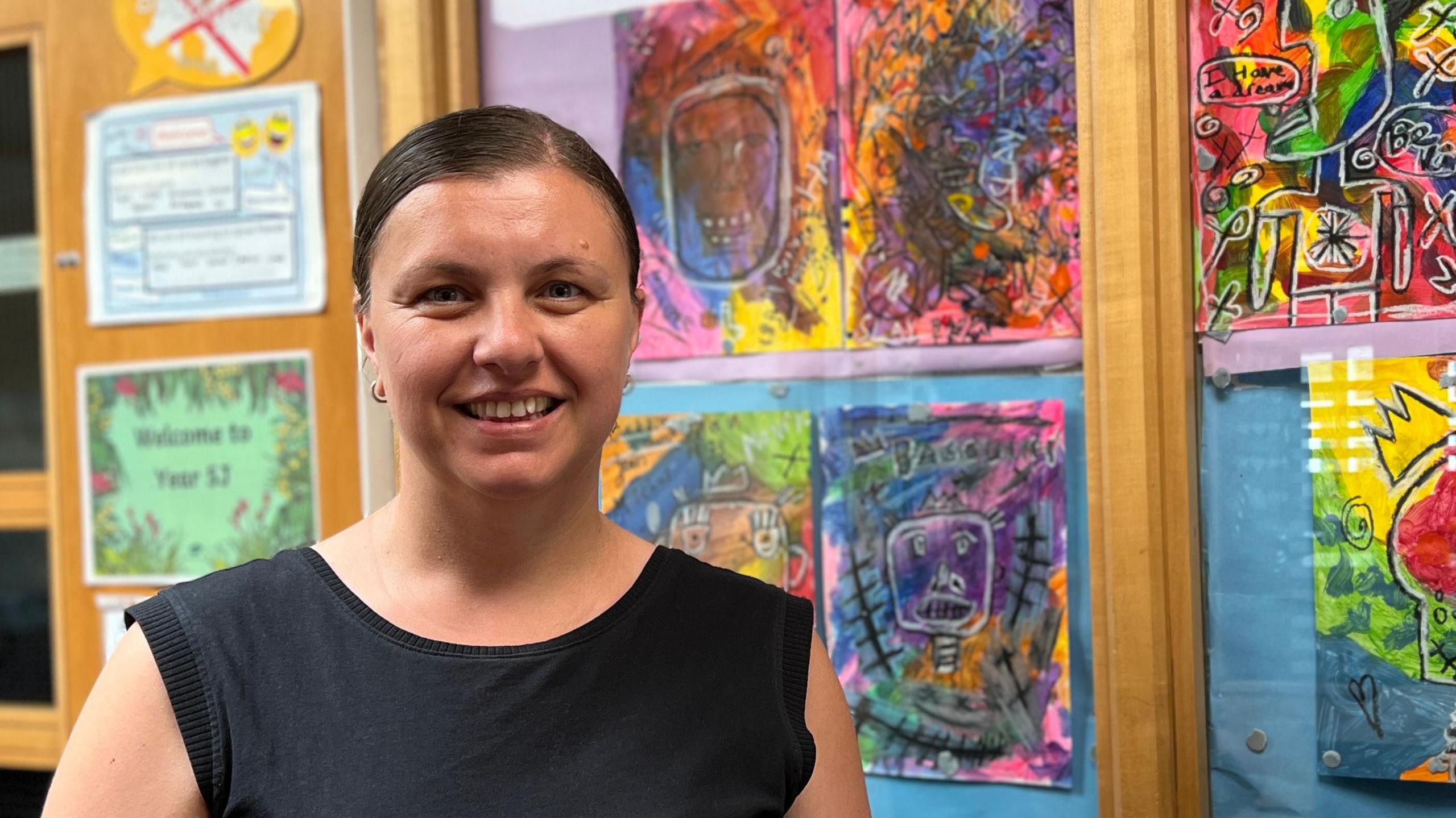 Georgiana is standing in front of a window decorated with colorful artwork and a 'Black History Month' sign. The window features vibrant, abstract designs. Nearby, a printer labeled 'Managed Print Service' with the number 874433 is visible. A door next to the window displays several signs, including one that reads 'Welcome Year 1.