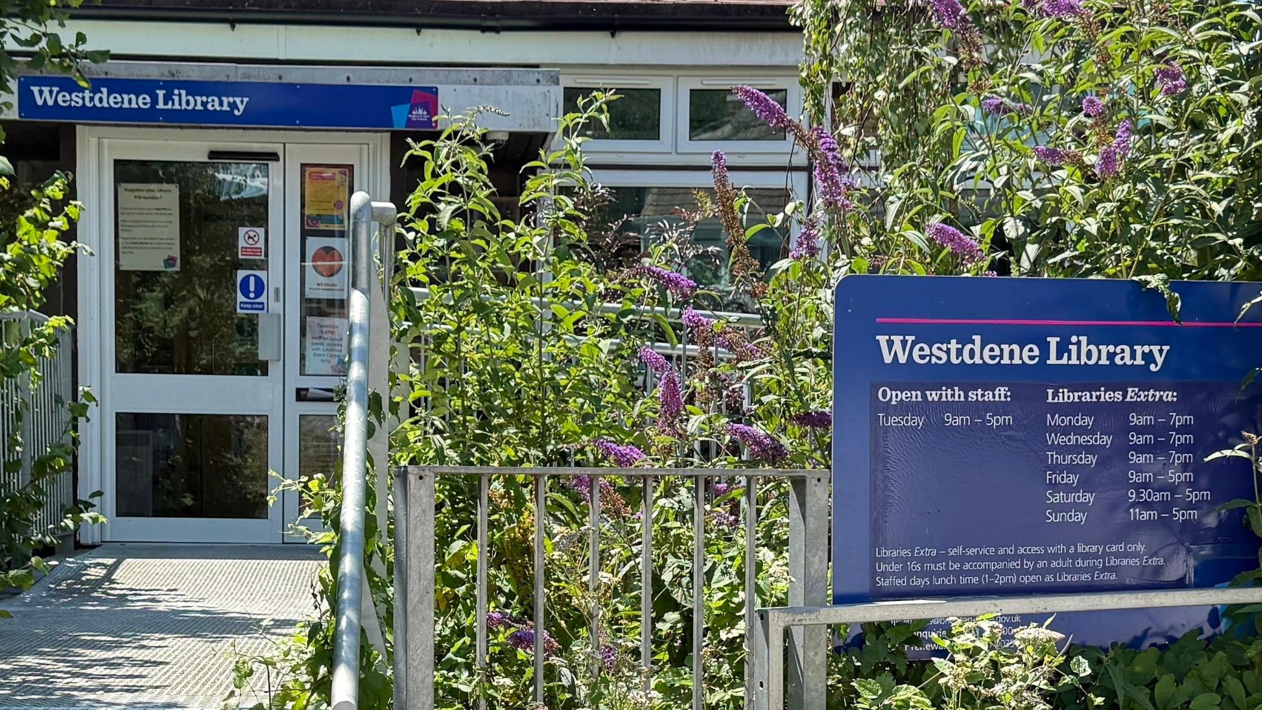 The image shows the outside of Westdene Library, with lavender planted outside a blue sign on the right, which has been placed behind a metal railing.
On the left-hand-side, the entrance can be seen, with another blue sign above the door reading "Westdene Library".