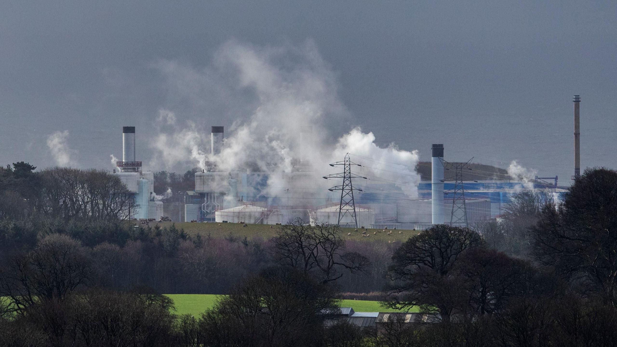 A wide view of the Sellafield site. Various grey industrial buildings can be seen on the skyline. Grey smoke is billowing from the buildings.