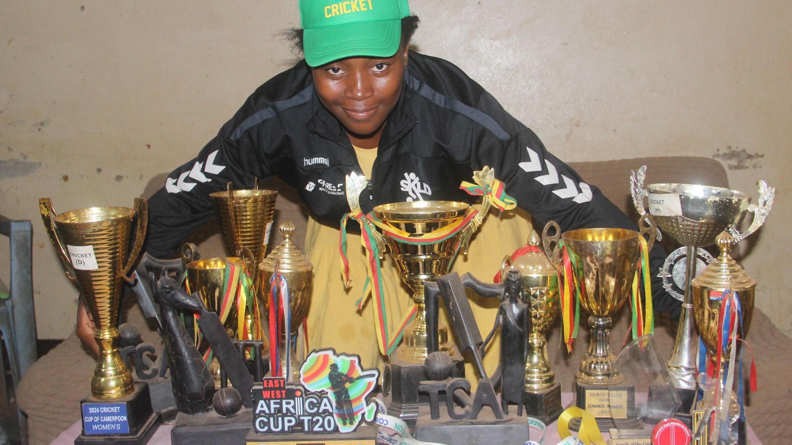 Madeleine Nseke Sissako, wearing a black tracksuit top over a yellow dress and a green baseball cap with the word cricket written on it in yellow, poses behind a table with a large collection of cricketing trophies, with some adorned with ribbons and one which says Africa Cup T20