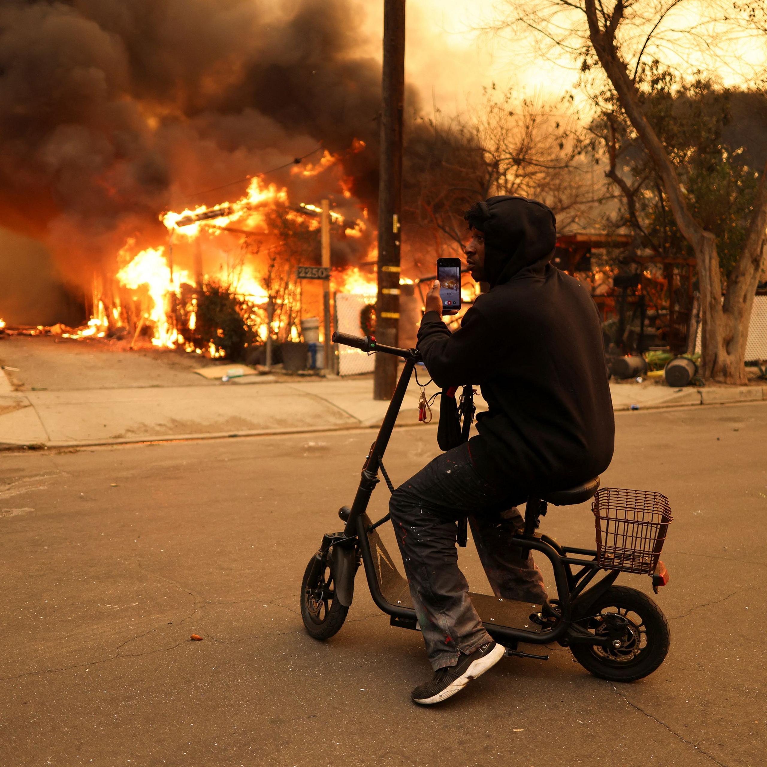 A person uses a cellphone to take a picture of a burning building while sitting on a stationary scooter with their hood up at the Eaton Fire in Altadena on Wednesday.