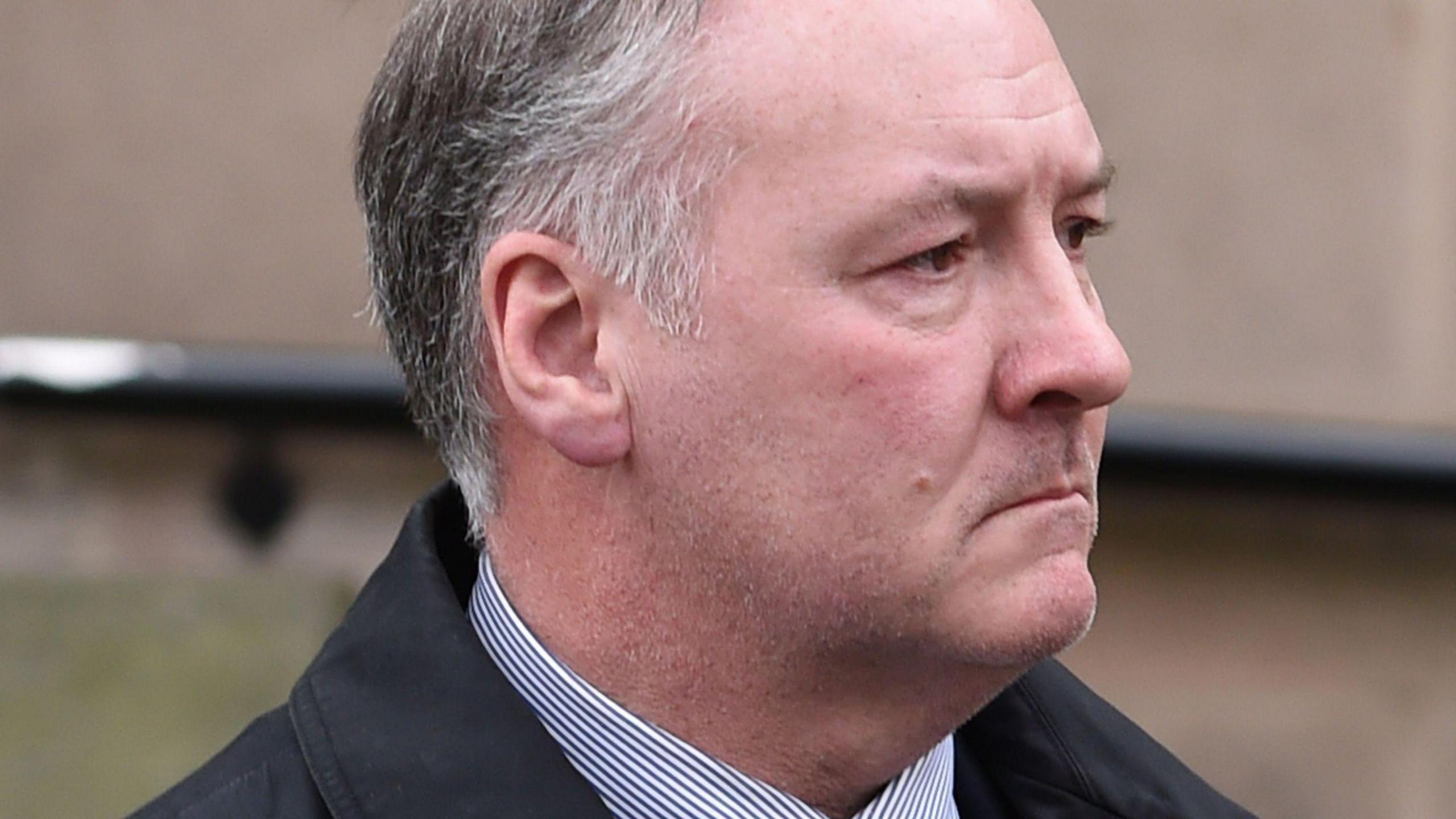 A close-up shot of Ian Paterson's face and neck as he arrives at court in 2017. Ian Paterson has grey hair, and is wearing a striped shirt and dark jacket. The background of the image is out of focus