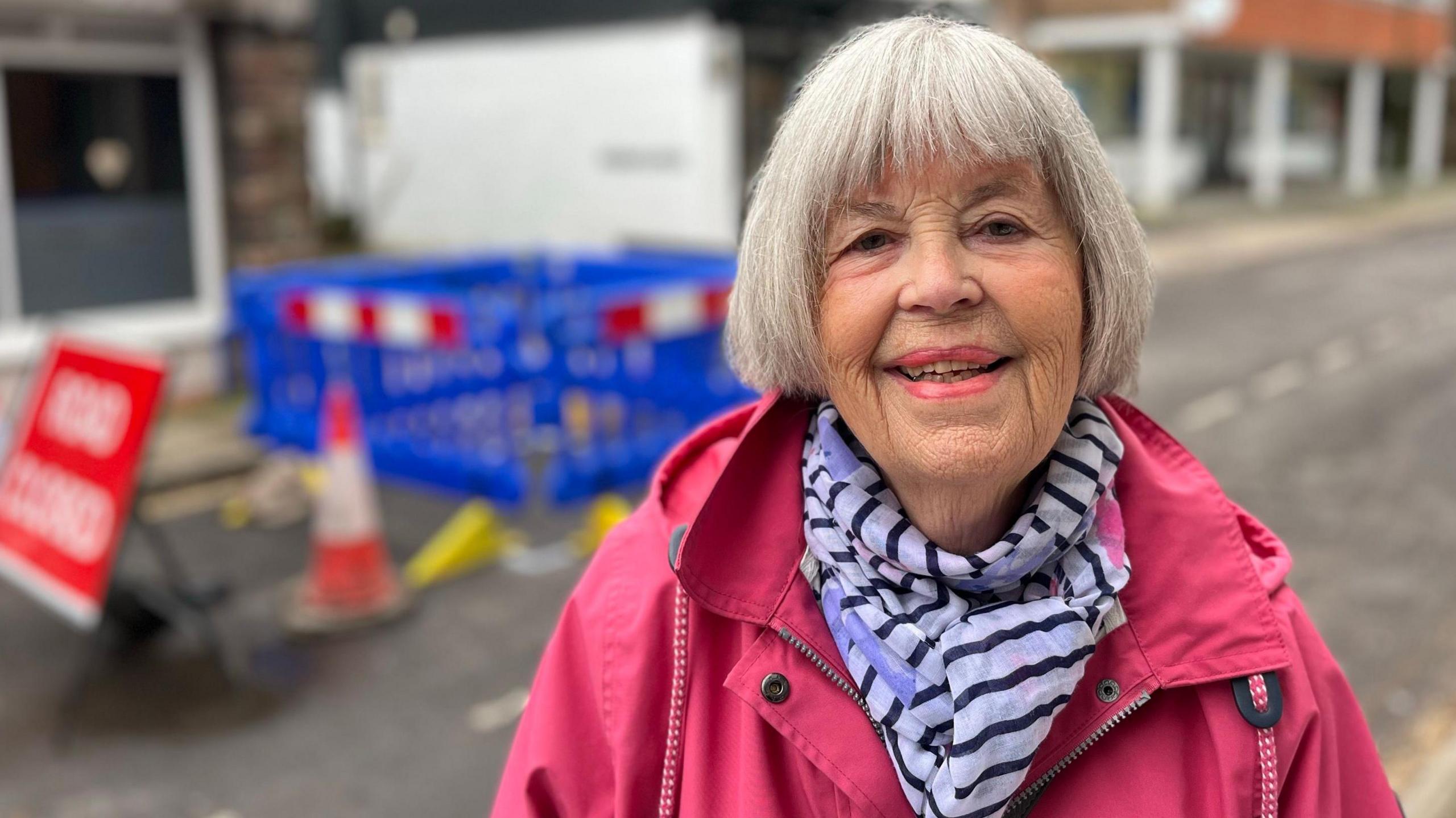 Hilary Fritche standing in front of the sinkhole wearing a bright pink jacket and a blue and white striped scarf. She has bright pink lipstick on and bobbed white hair. She is smiling