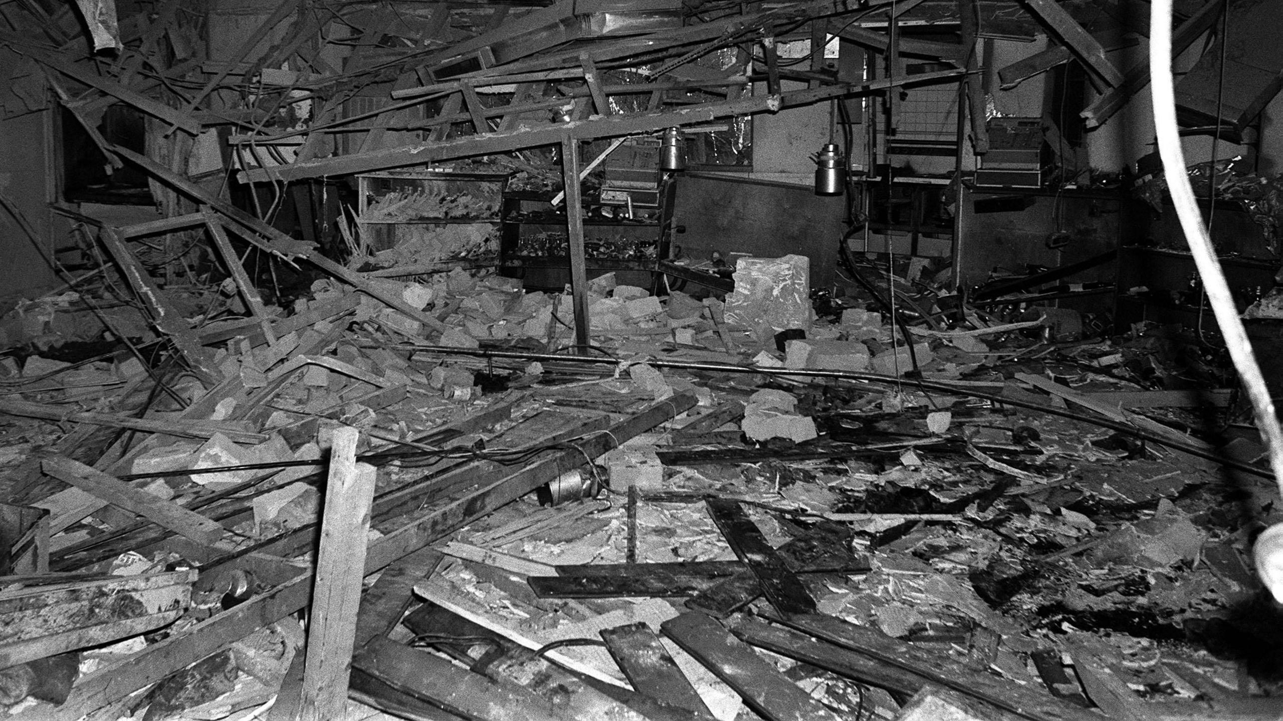 A black and white image showing a destroyed room interior. Metal beams and rafters hand down, warped, bent and broken. Planks, insulation and rubble can be seen on the floor.