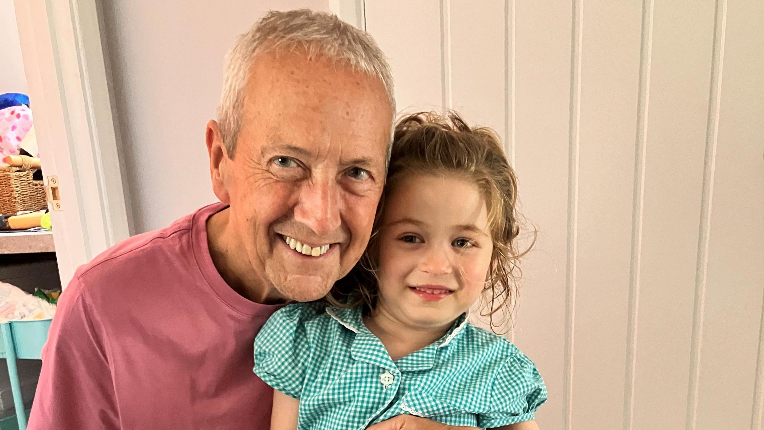 Stan, who has short grey hair and wears a light red t-shirt, smiles as he holds Maggie while they pose for a picture. She is wearing a green and white checked summer school dress, and has her blonde wavy hair tied back in a ponytail.