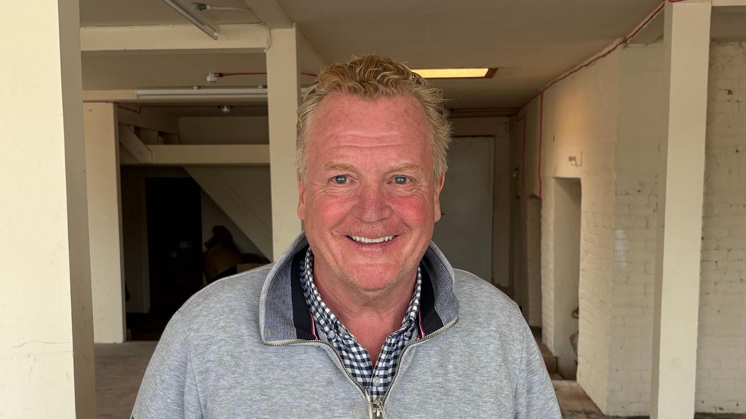 A man with greying red hair wearing a checked shirt and grey quarter-zip top standing in a derelict room.