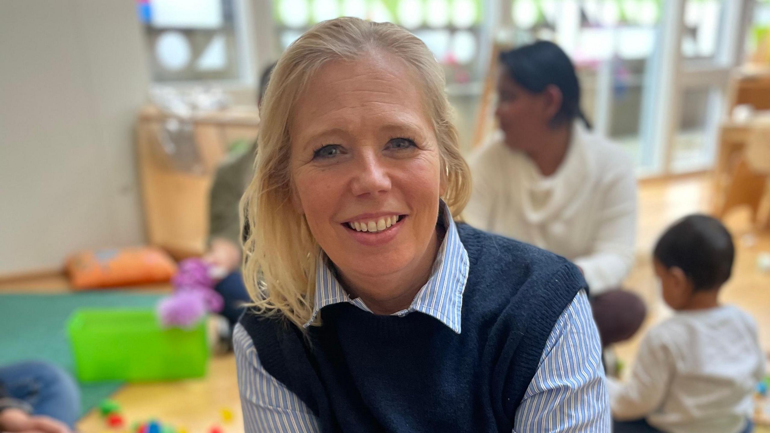 Fiona is sat crossed-legged on the floor of the nursery. She is smiling directly at the camera. Behind her - out of focus - a mother and children are also sat on floor playing games. There are toys strewn across the floor. Fiona is wearing a blue and white striped shirt and a dark blue tank top sweater.