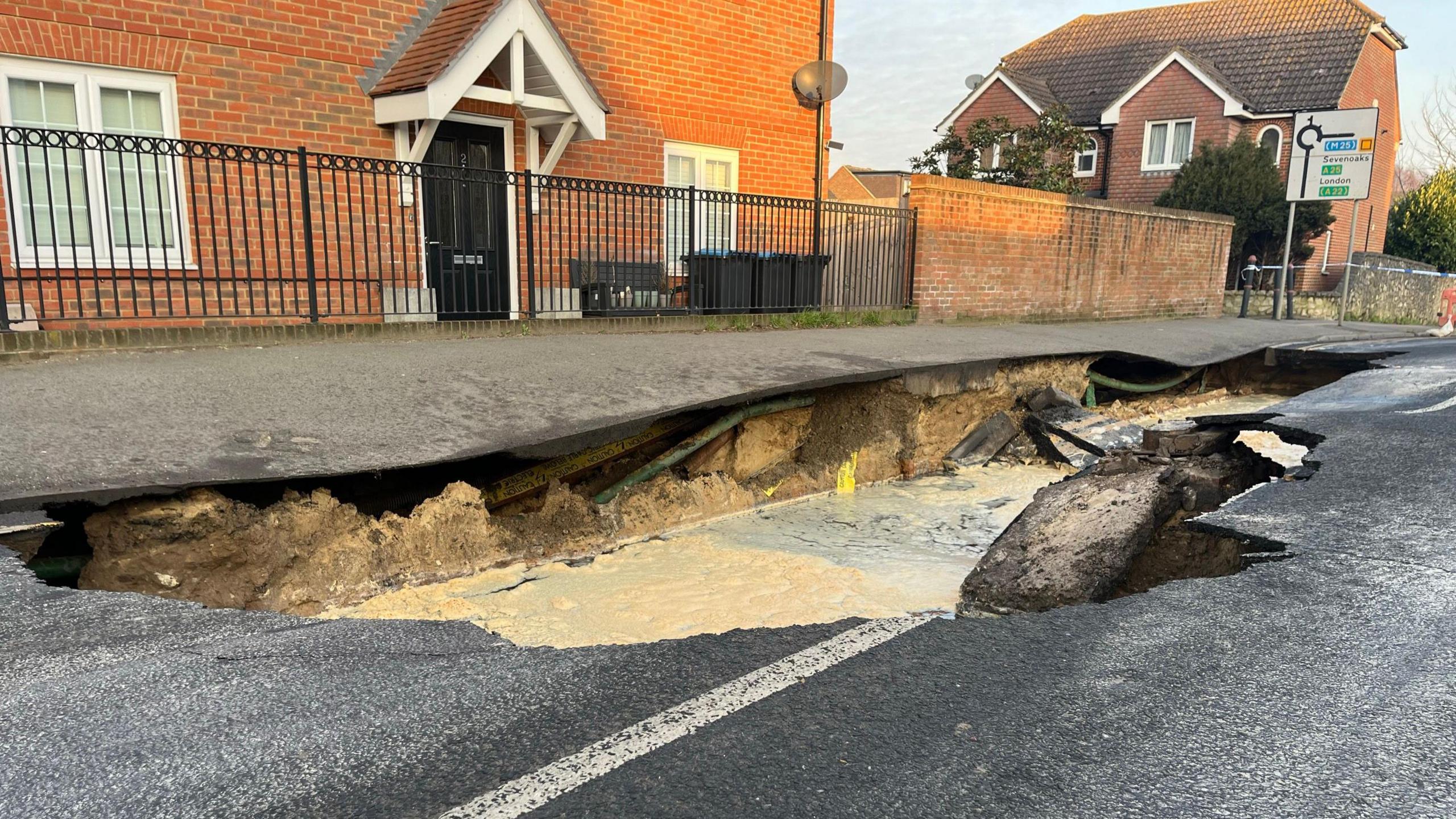 A sinkhole which has caused a large amount of damage to Godstone High Street. There is a house in the background, as well as a road sign. 