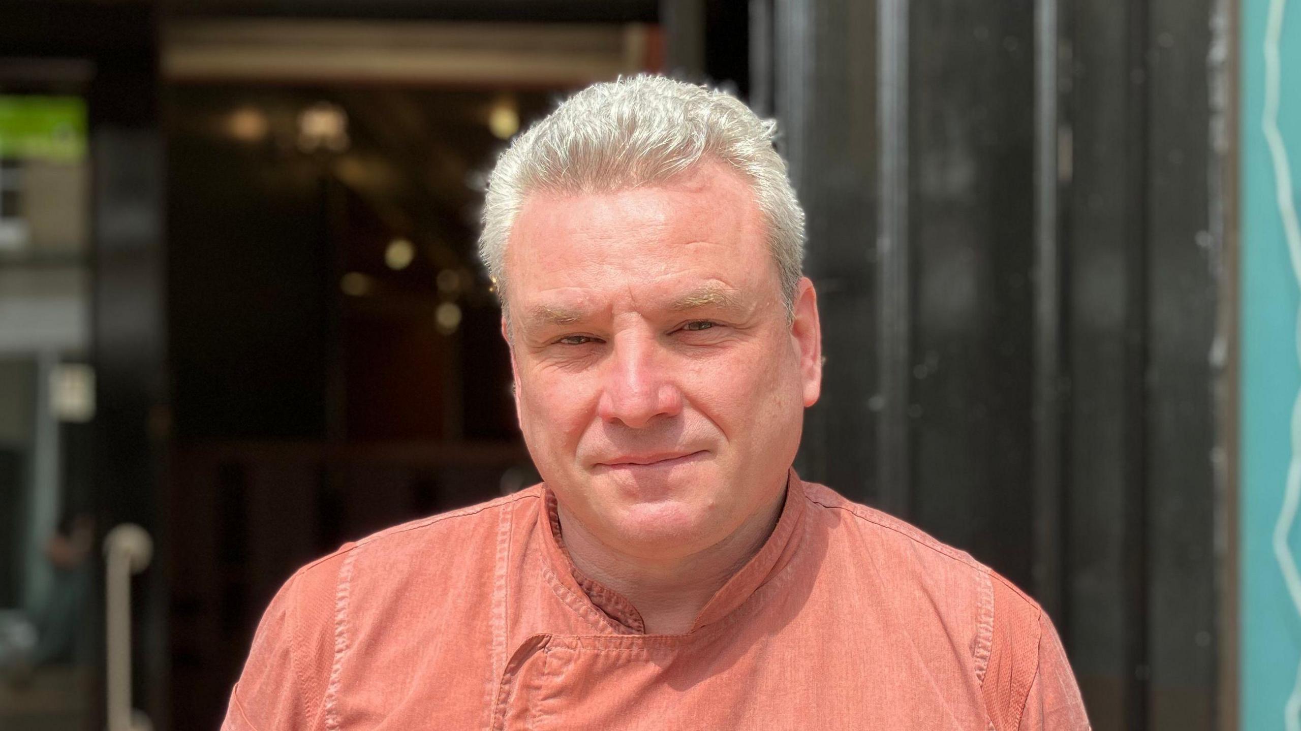 Stuart Calder, a middle-aged man with short grey hair and glasses, is pictured standing in the doorway of a cafe. He is wearing a buttoned orange shirt. Behind him the door can be seen which is slightly blurred.