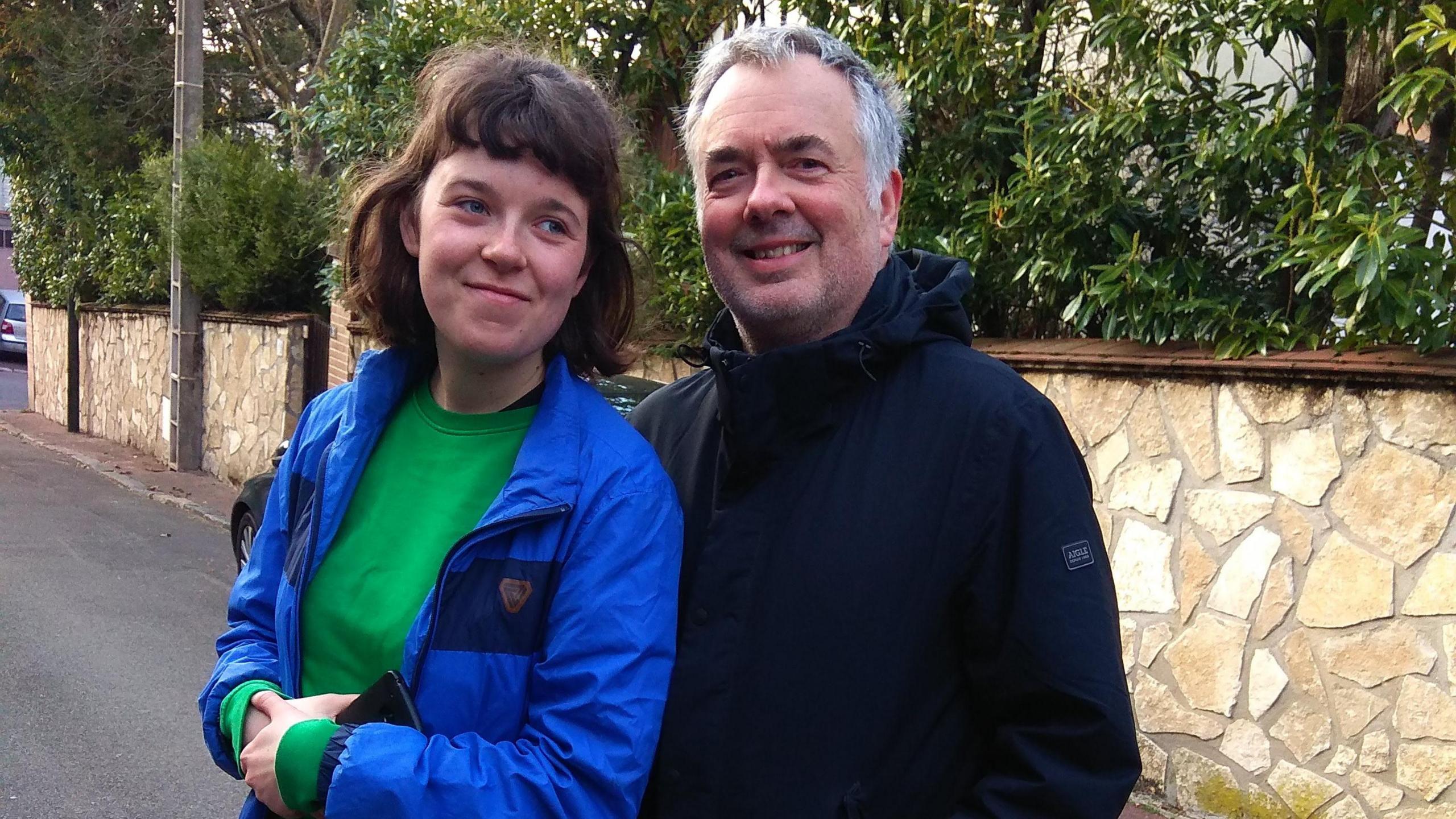 A young woman pictured standing close to her father. She is wearing a long-sleeved green round neck top and a blur jacket. Her hands are clasped and she is holding a mobile phone. She is looking off to the left and not directly at the camera. The man, who is in his 60s and has short grey hair, is wearing a black hooded jacket which he has zipped up to the neck. He is smiling and looking directly at the camera.