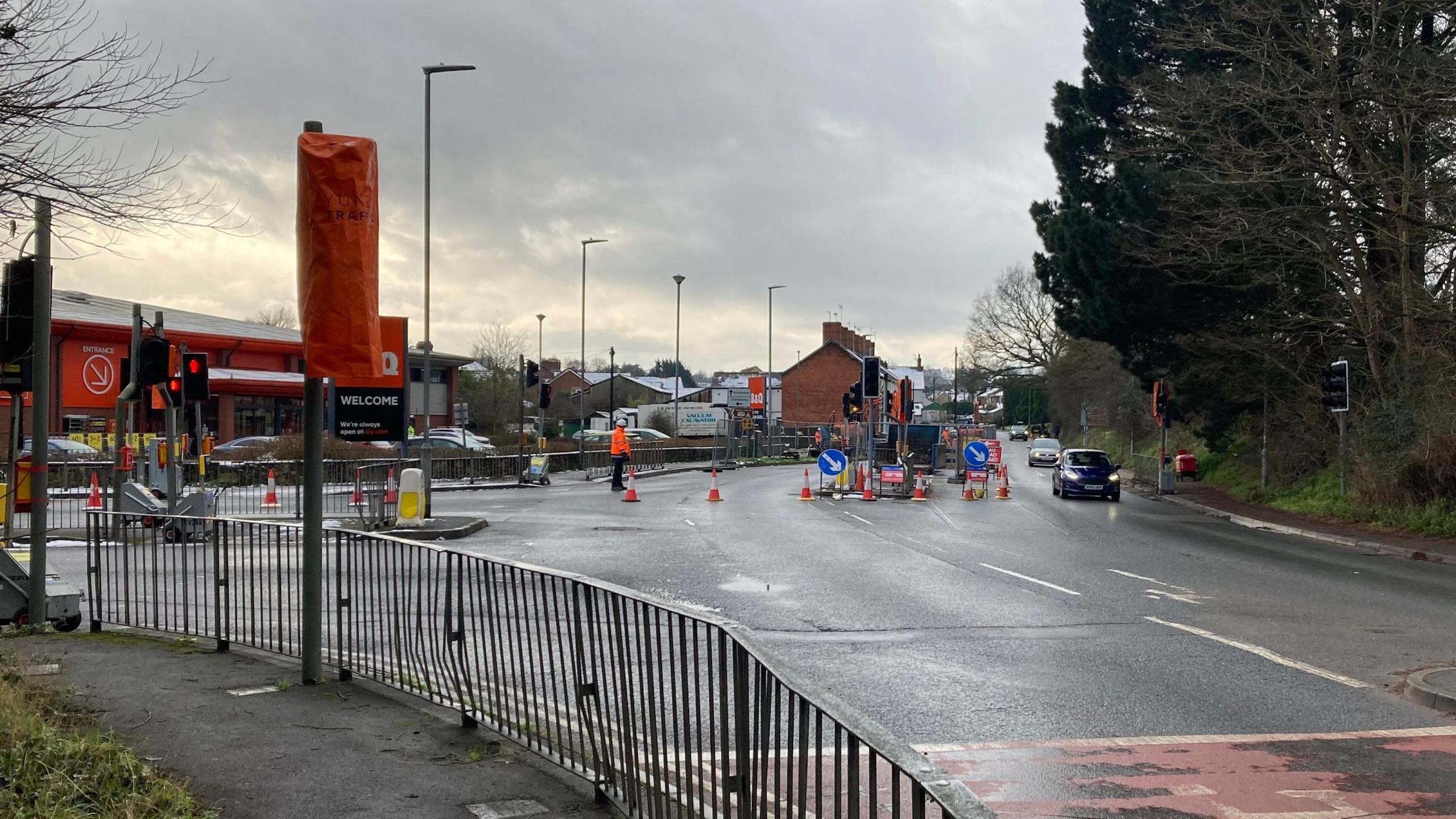 Roadworks and temporary traffic lights in front of a zebra crossing. There's a house in the background with road work barriers and cones in the foreground. There's a B&Q shop and queues of cars.