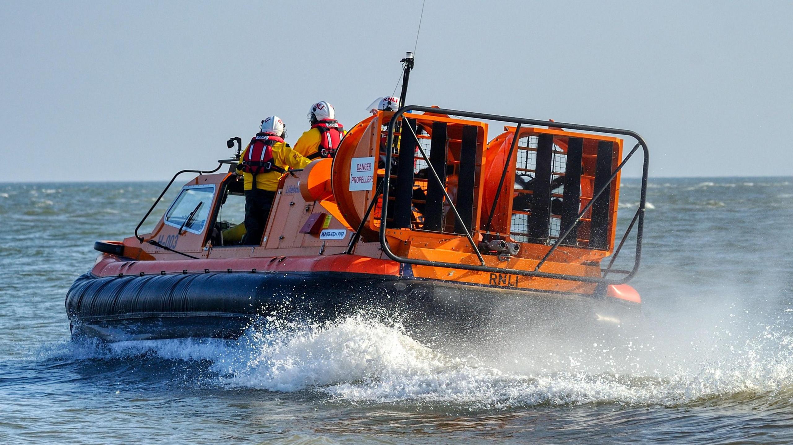 An orange hovercraft is on the water heading away from the camera. On the back, it has two hovercraft engines and a sign which says RNLI.