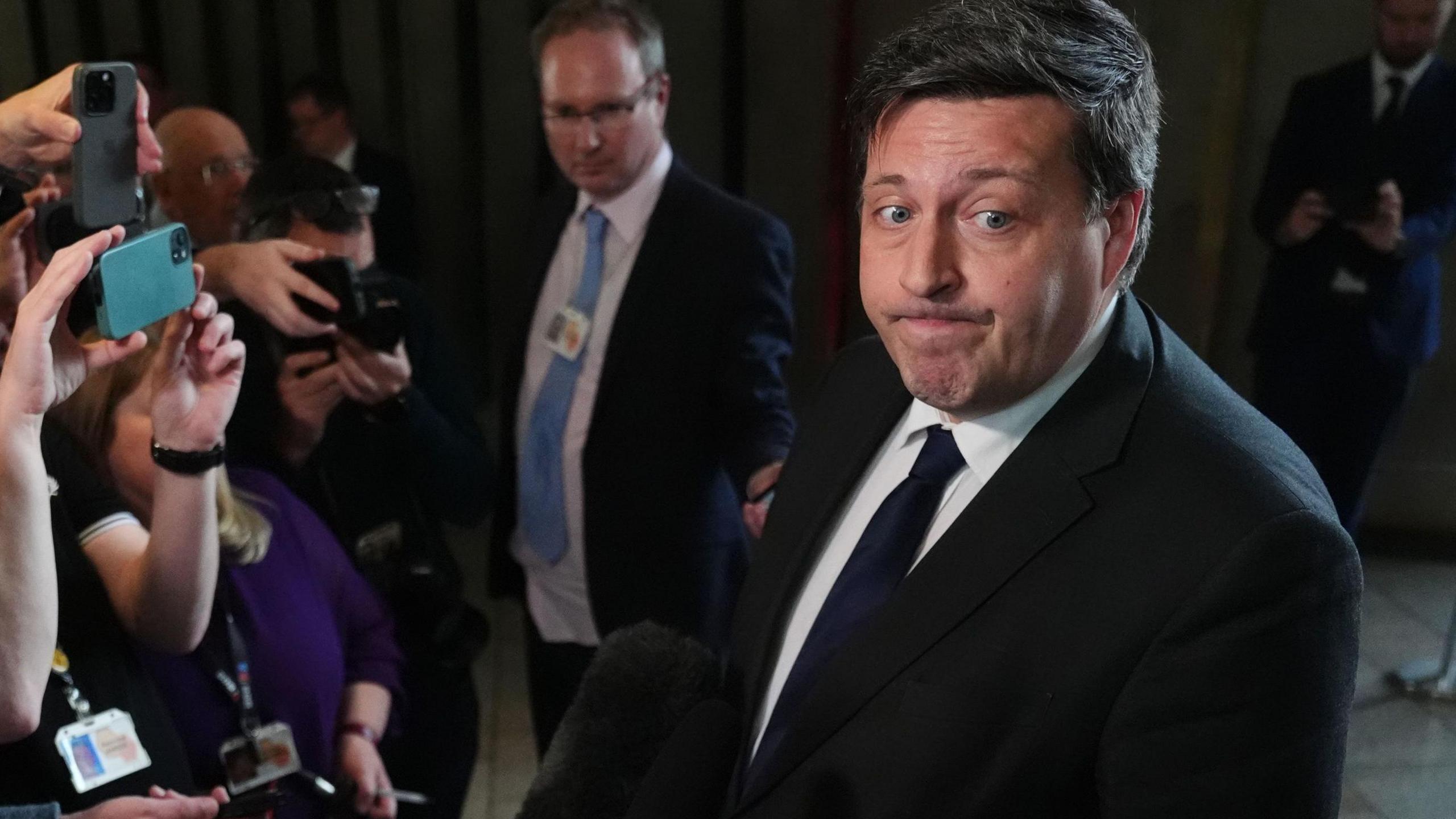 Jamie Hepburn looking off camera. He is wearing a dark suit jacket over a white shirt with a dark tie. Several cameras and mobile phones are being held up to him.