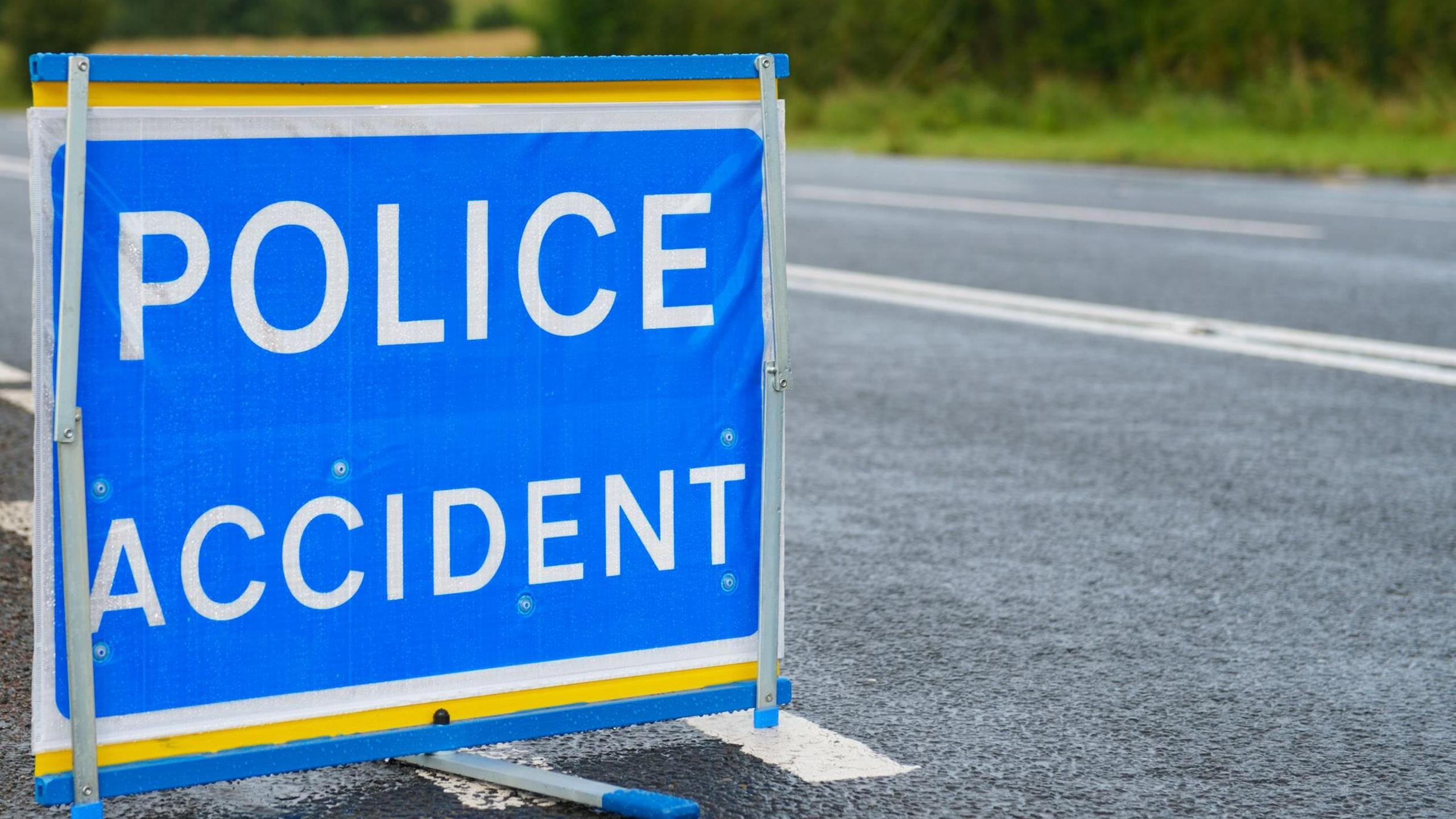 A square blue sign saying police accident in white letters. It is sitting on a tarmacked road. Beyond the road can be seen a grassy verge and shrubs.