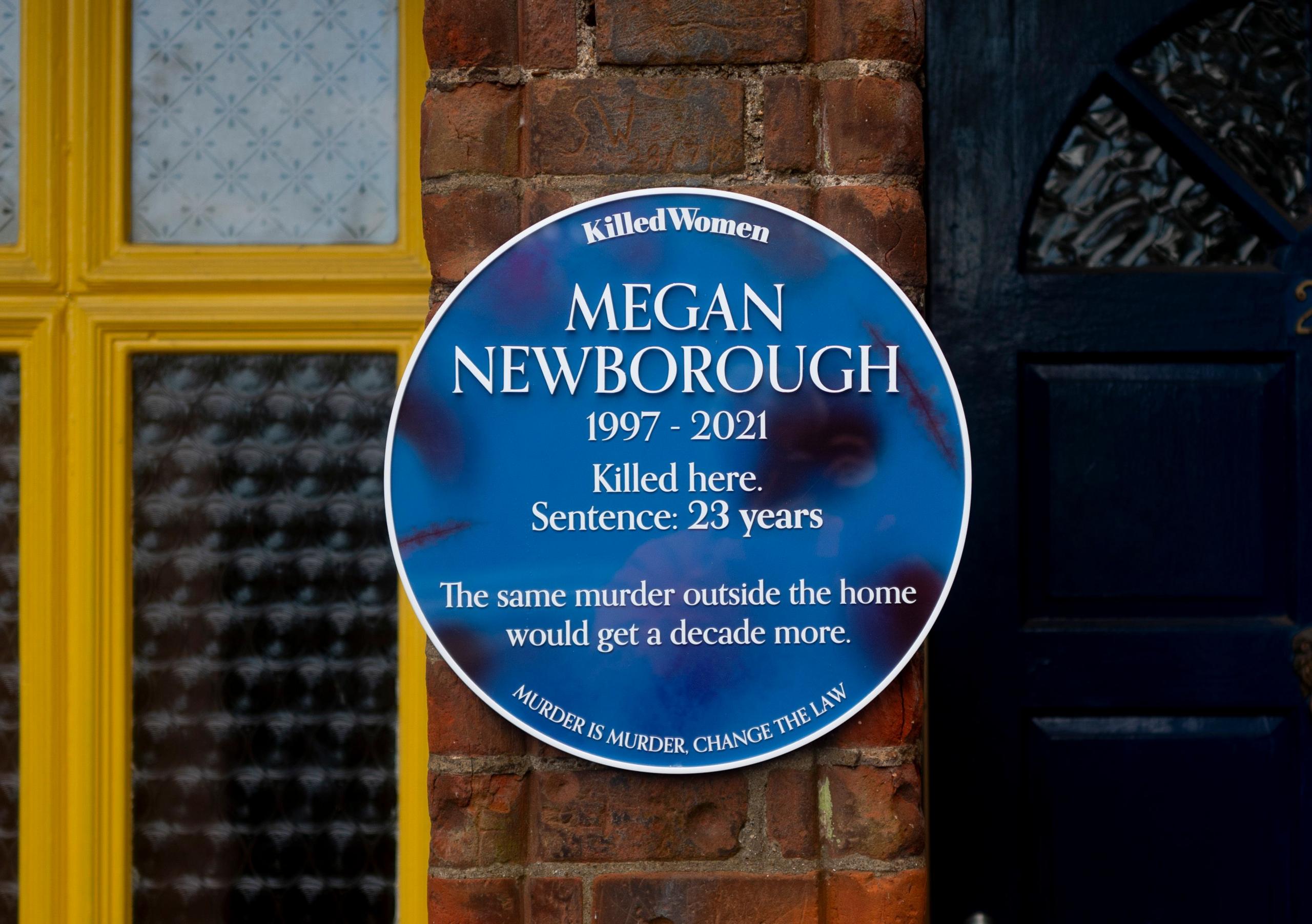 A blue plaque with black marks which resemble bruising is attached to a brick wall in between a black door and a yellow-framed window.