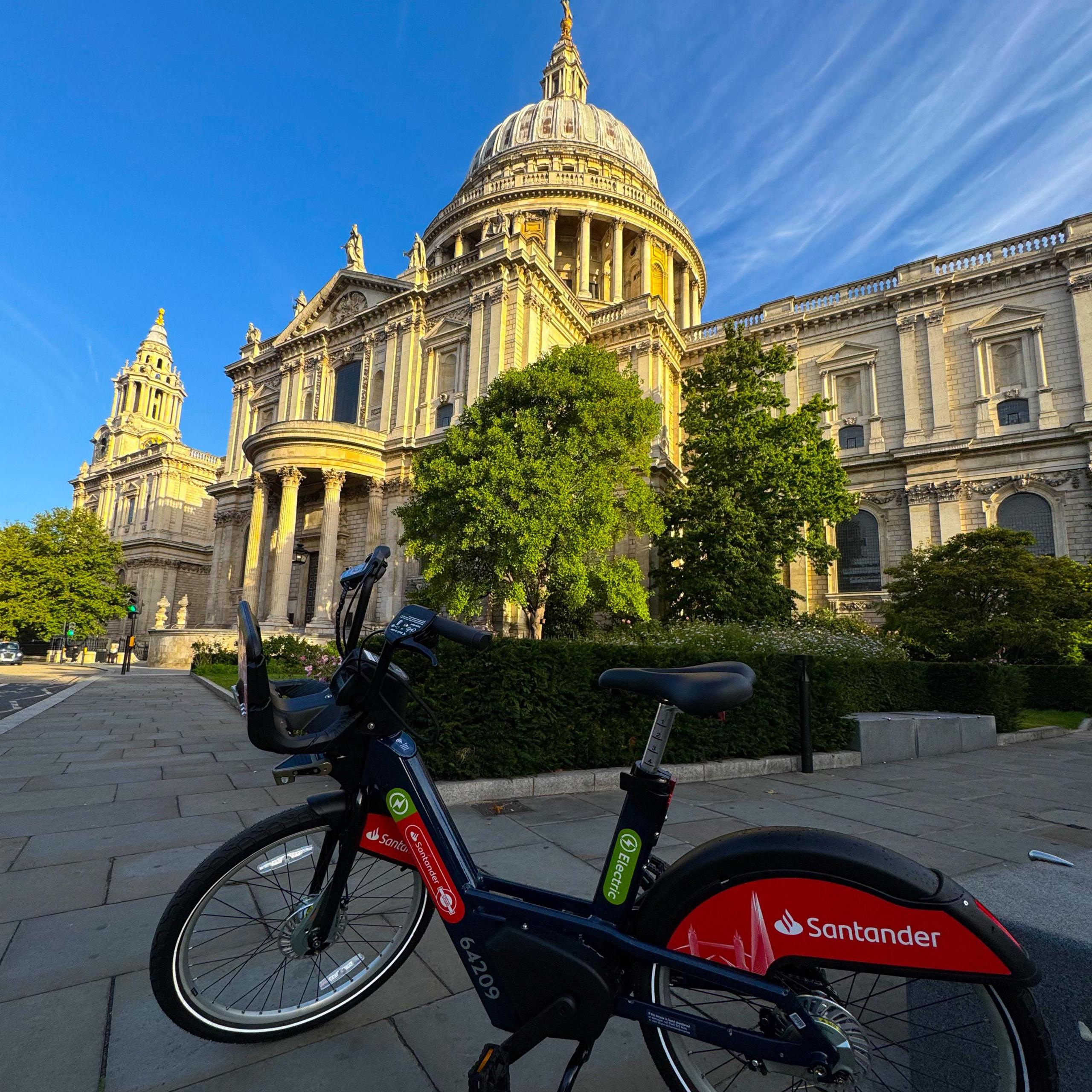 An electric Santander Cycle with red branding is parked on a wide pavement in front of St Paul’s Cathedral. The dome and towers of the cathedral rise above, with green trees partly visible against a bright blue sky.