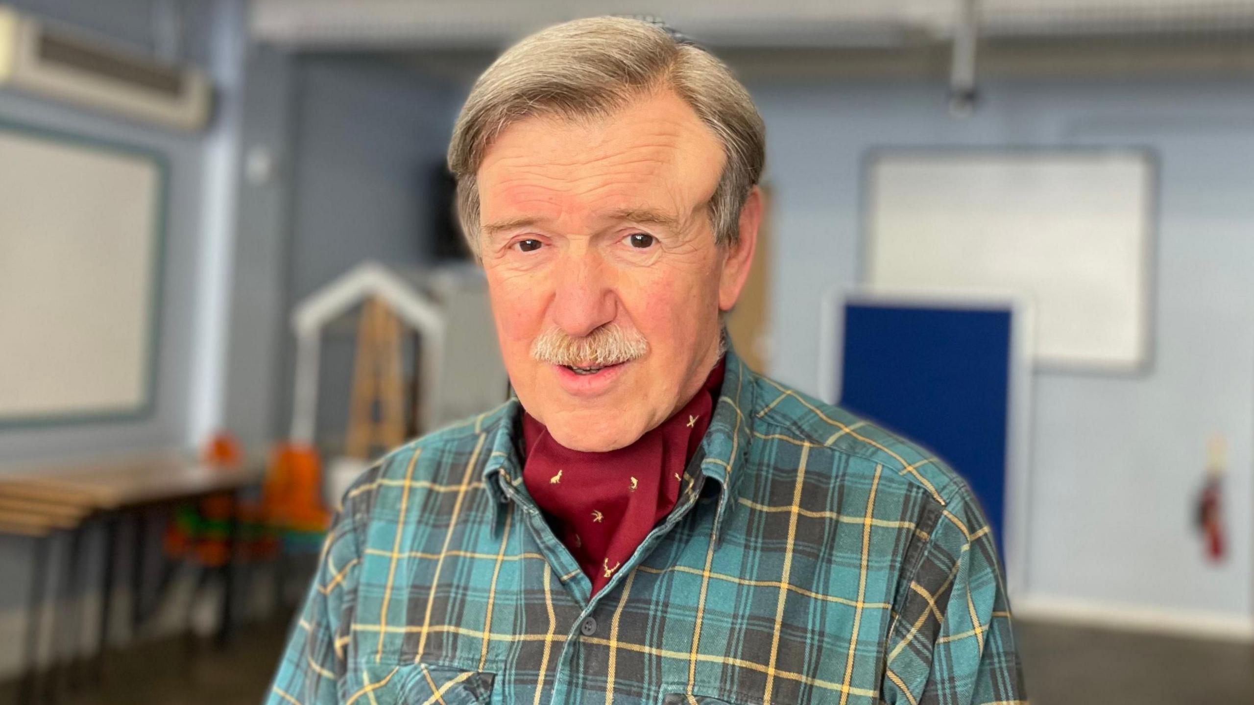 Stuart Prochnow looking at the camera, with a yellow and blue checked shirt. In the background is a community meeting room.