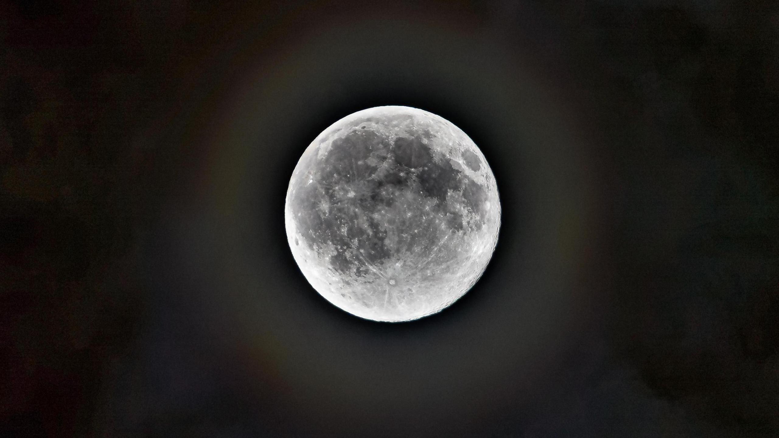 A close up of the full moon. It is a bright white colour and you can see the craters in grey. There is halo around the moon against the black sky.