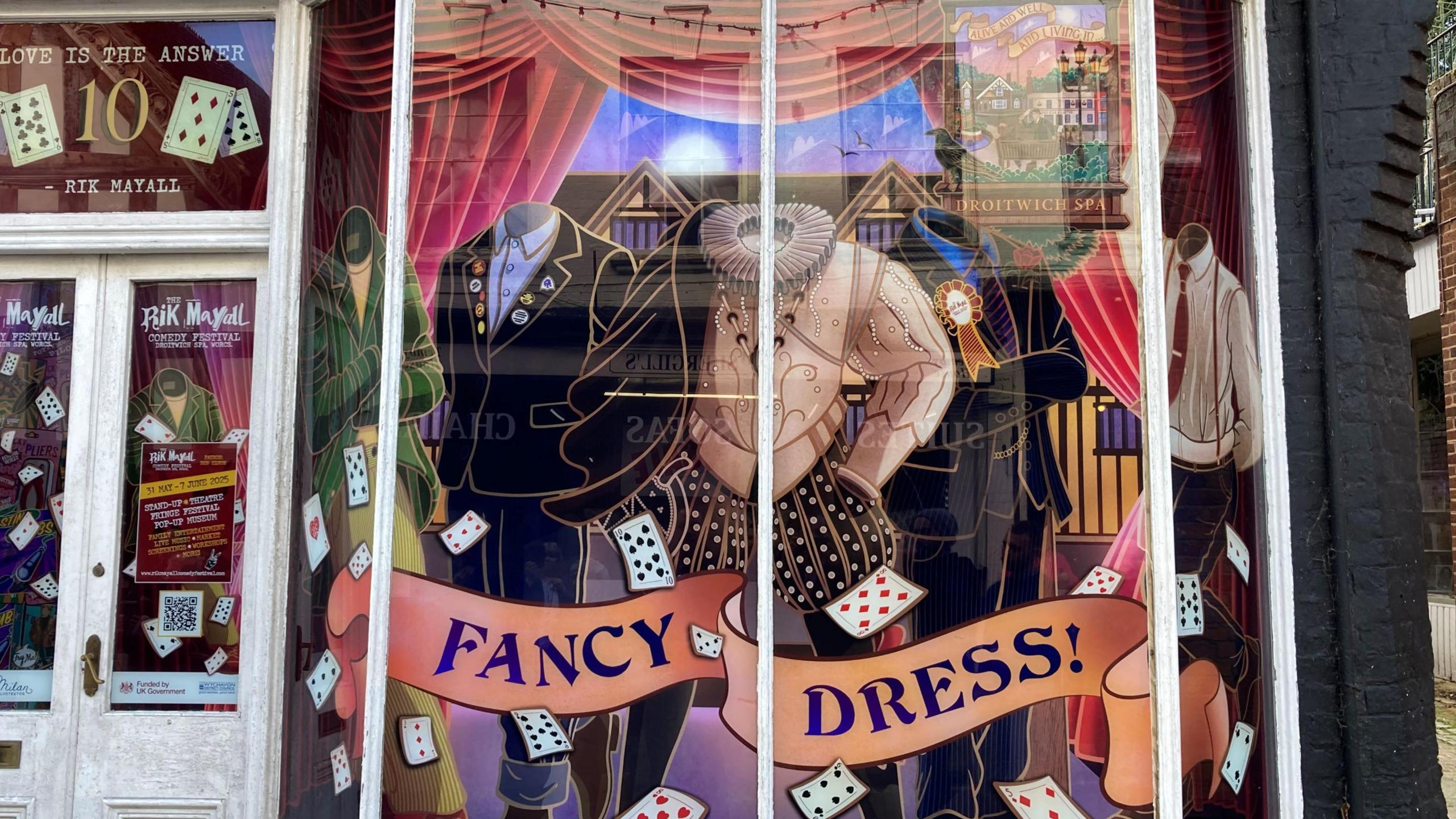 A painted mural in an old shop window features several of Rik Mayall's famous character outfits, floating playing cards and the words "Fancy Dress" written across a peach banner. 