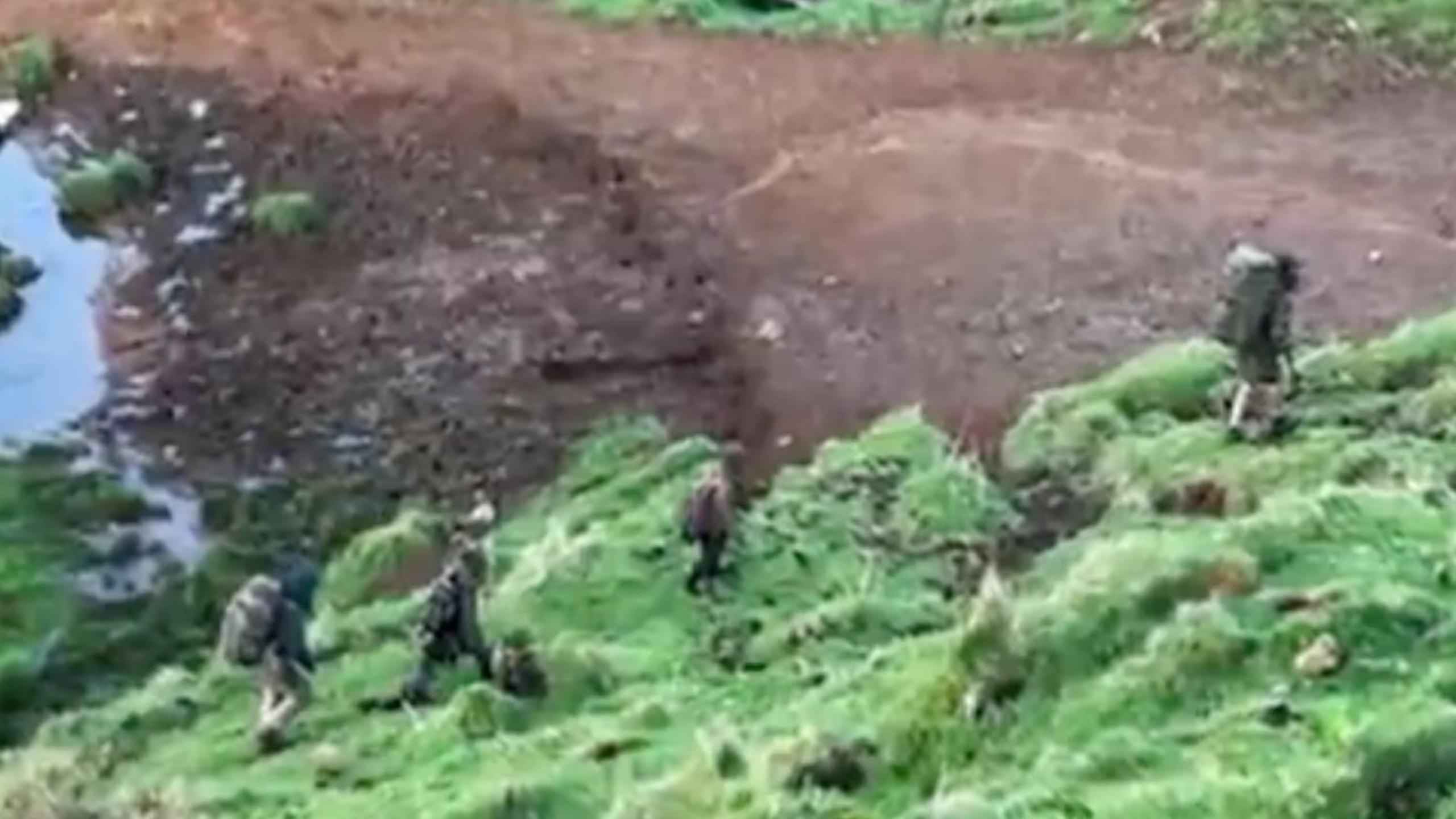 A blurry screenshot of a video taken on a phone showing four people pictured from above walking in the wilderness carrying rucksacks and wearing outdoor gear, in October 2024.