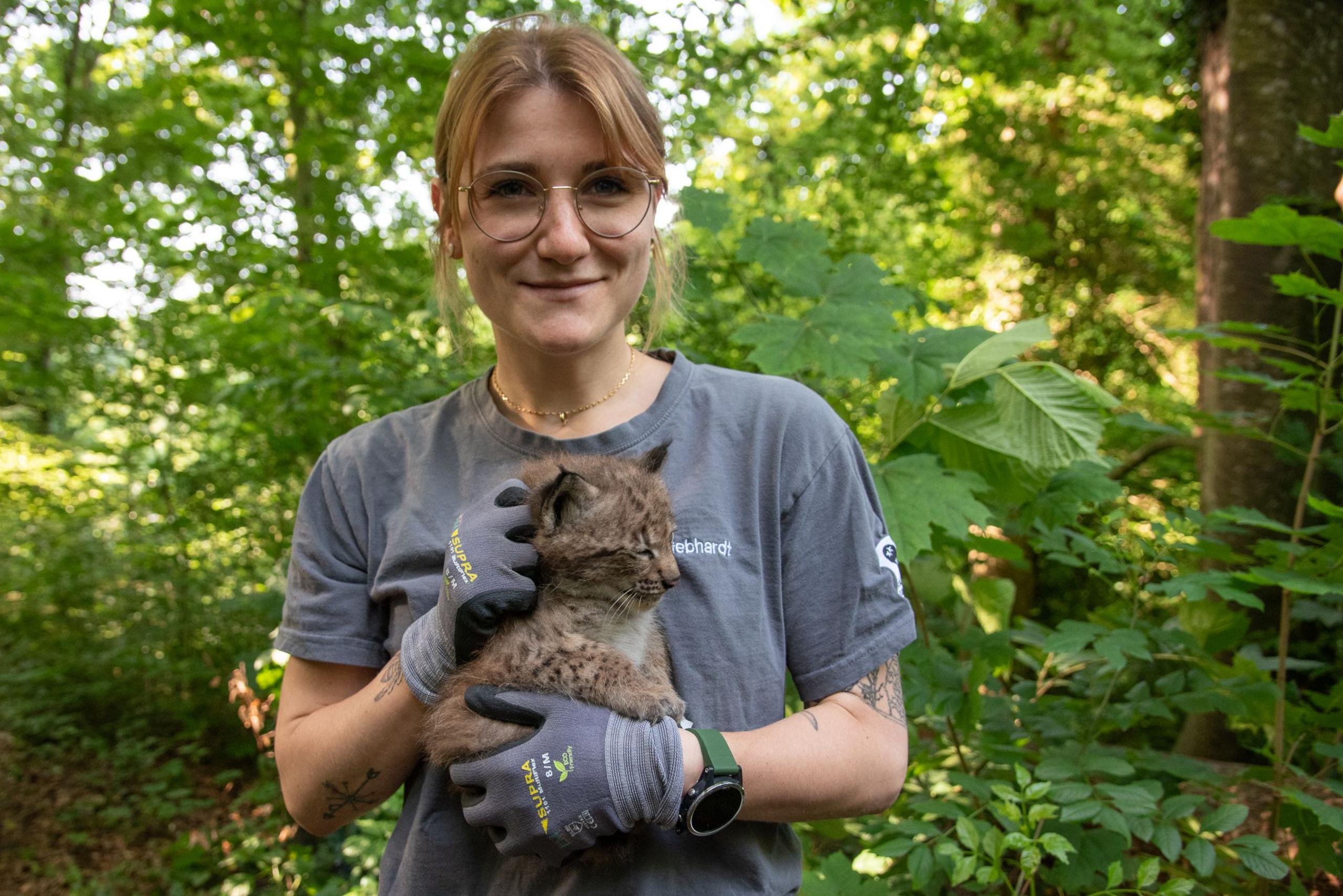 Dina Gebhardt wearing glasses is holding a small baby lynx kitten in her hands and smiling. She is standing in front of what appears to be forest with a smile on her face.