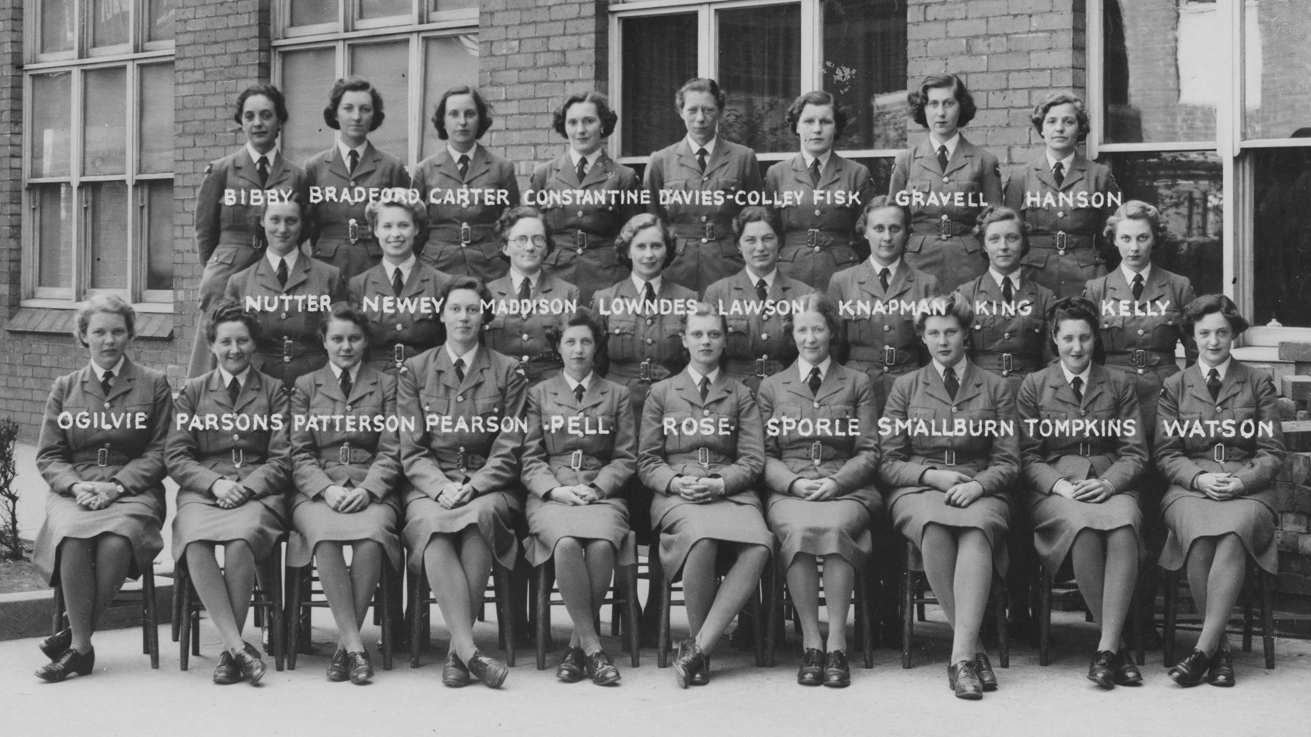 A posed portrait of about 25 women sitting outside a bricked building. The front row are sitting, and the middle and back rows appear to be standing. They are in military uniform. It is black and white.