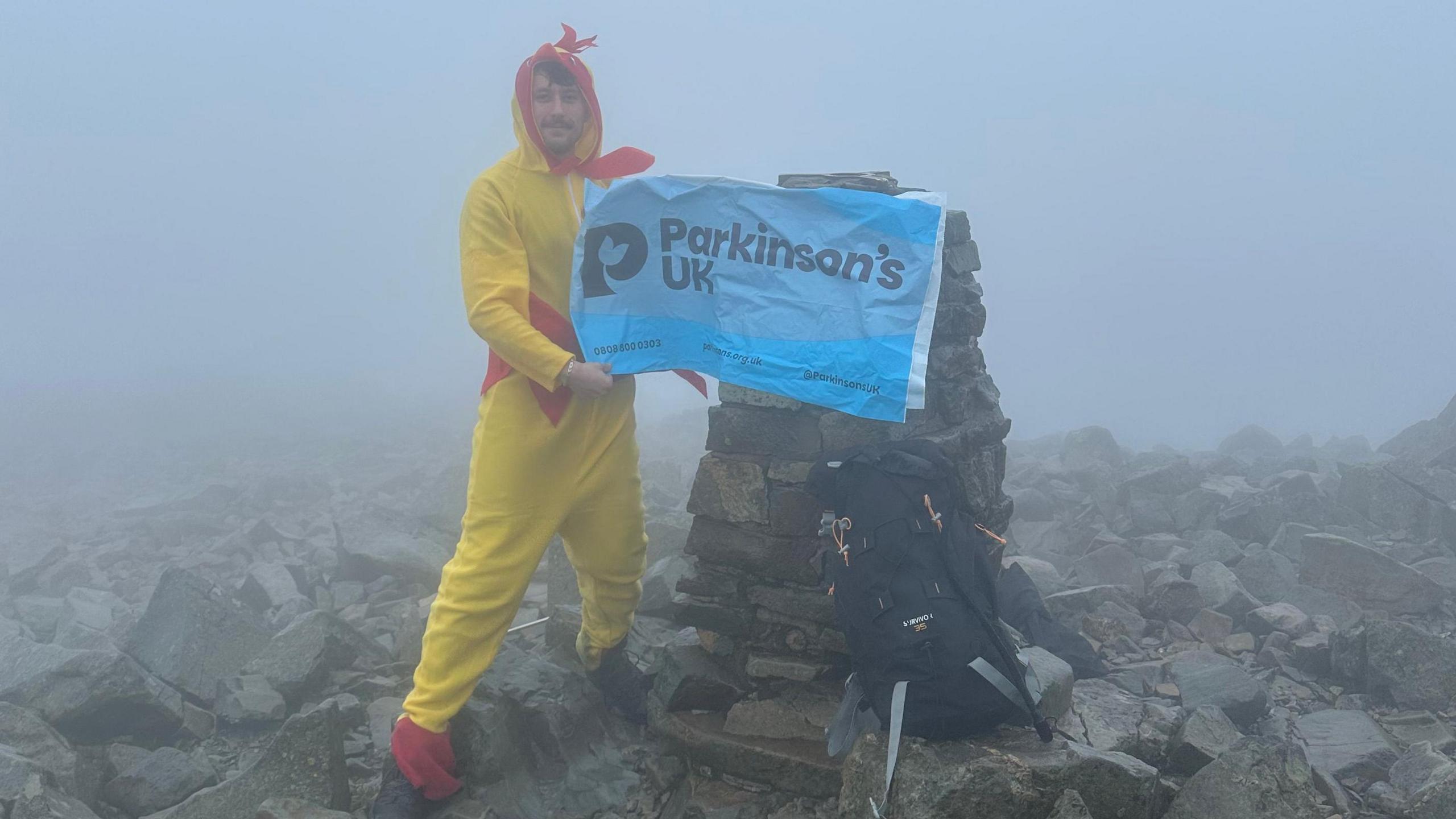 Luke unfurls a Parkinson's UK flag at the top of the mountain. The surface is rocky, and there is thick fog.