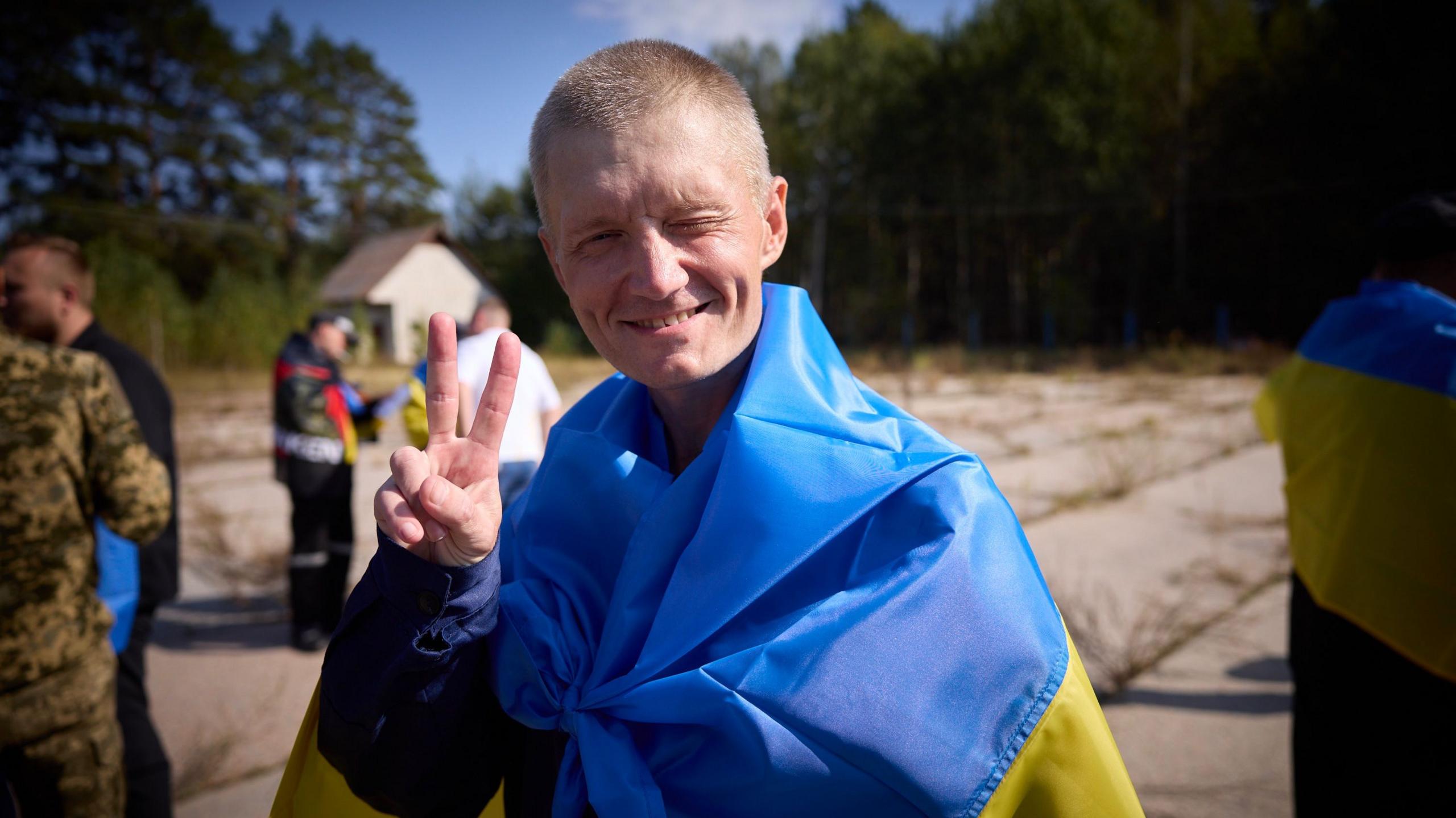 A Ukrainian servicemen released during a prisoner exchange with Russia is seen squinting and smiling into the camera. He is also wrapped in a Ukrainian flag and holding up two fingers facing forward in a V sign. 