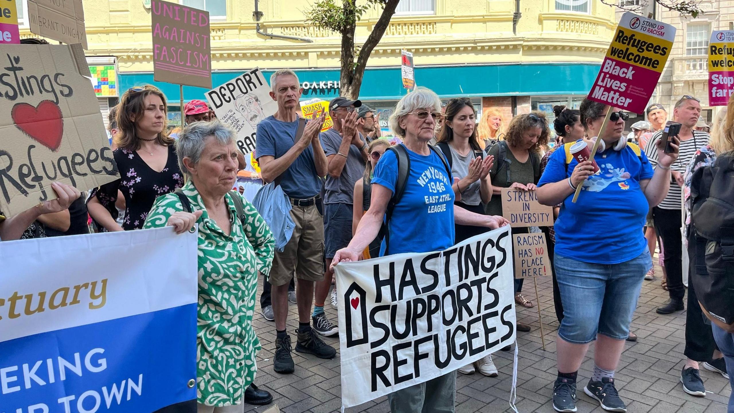 A crowd of mostly women can be seen holding banners in a Hastings street which say slogans such as "Hastings Supports Refugees"