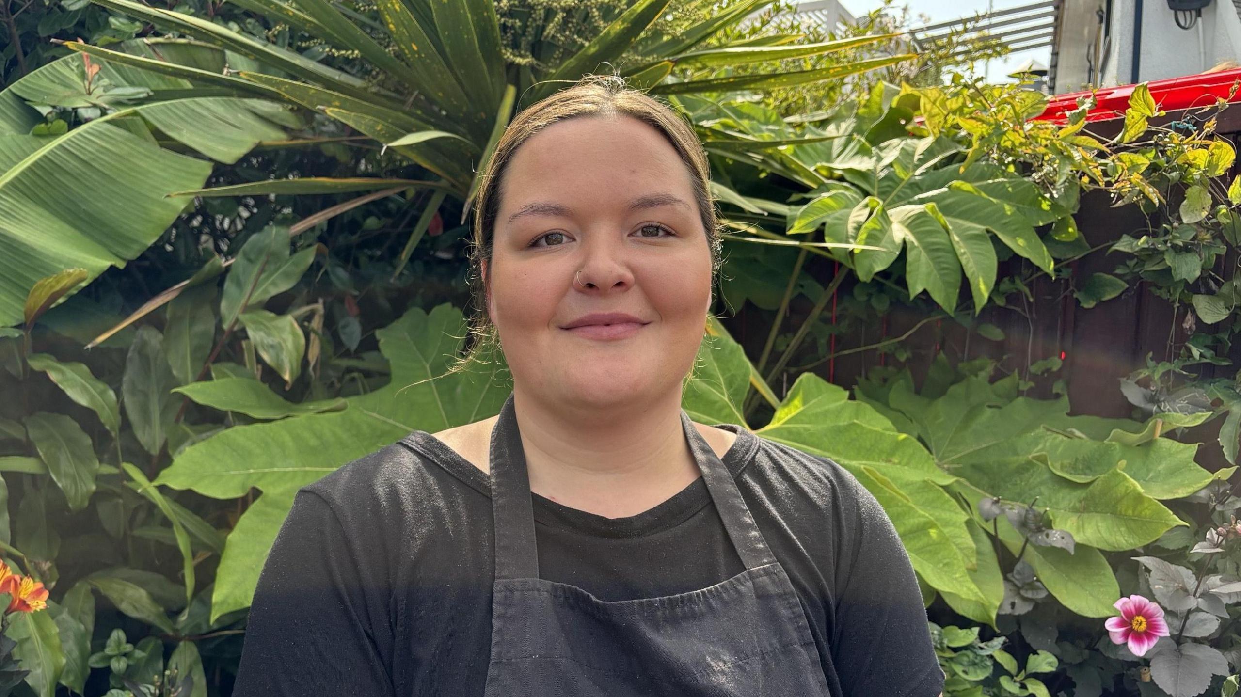 Megan Holyoak in black t-shirt and black apron looking at the camera, she is standing in front of some greenery