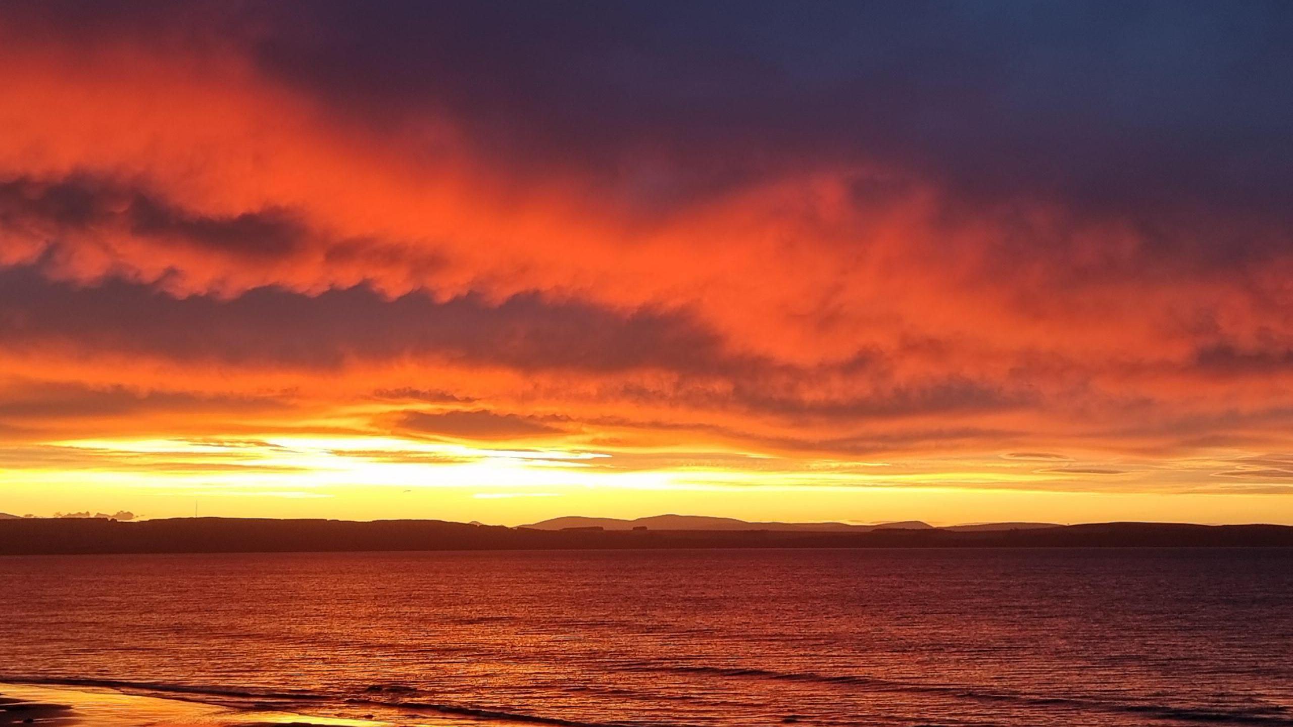 View across the sea to a mountain range on the other side on the horizon.  Sky is filled with cloud and with a red and orange tinge of the setting sun.