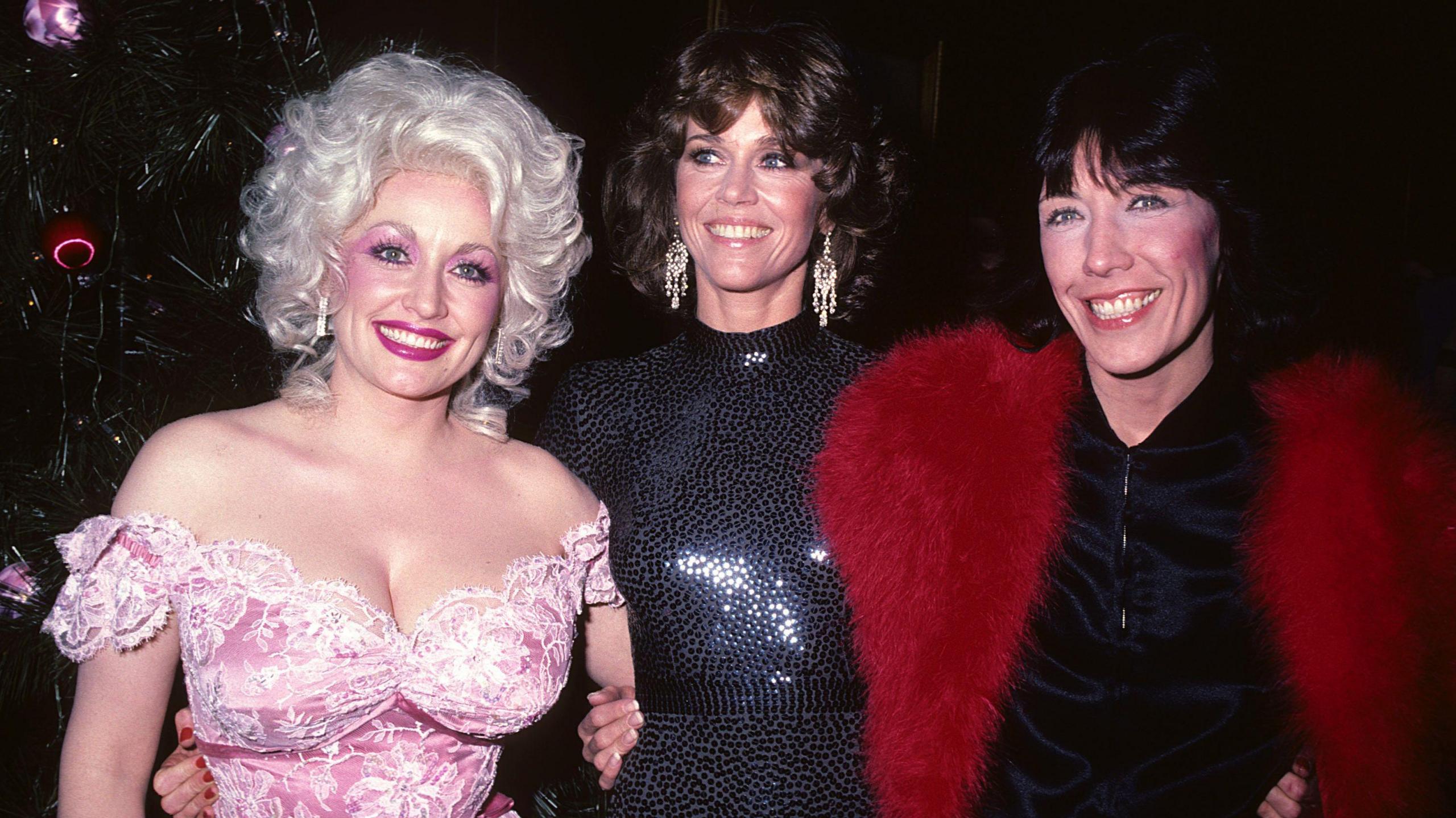 Dolly Parton, wearing a bright pink off-the-shoulder dress, Jane Fonda in a black sequinned high neck dress, and Lily Tomlin in a black dress and bright red fur wrap.