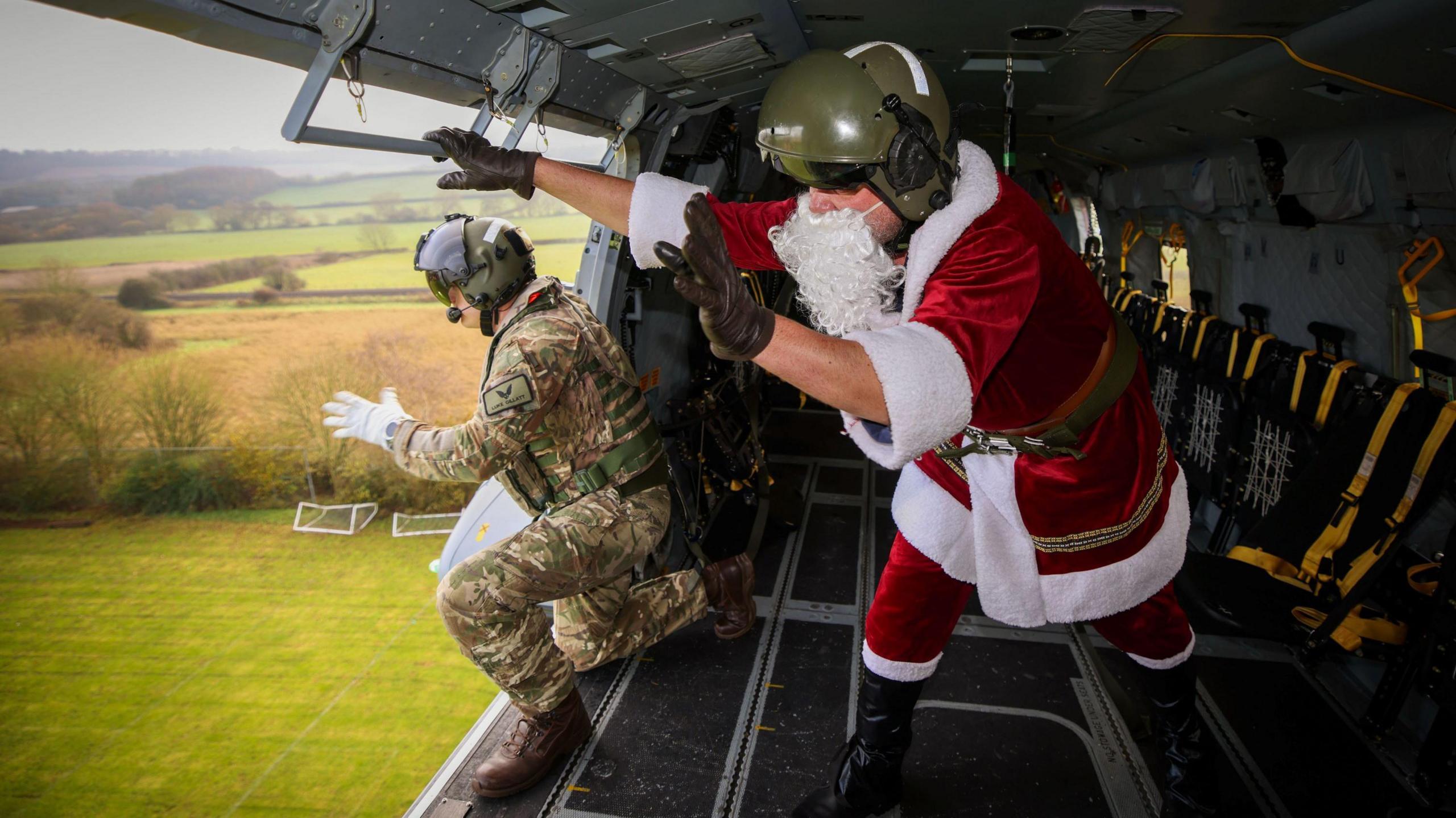 Father Christmas and a man in camoflage gear inside a helicopter that is just above the ground.