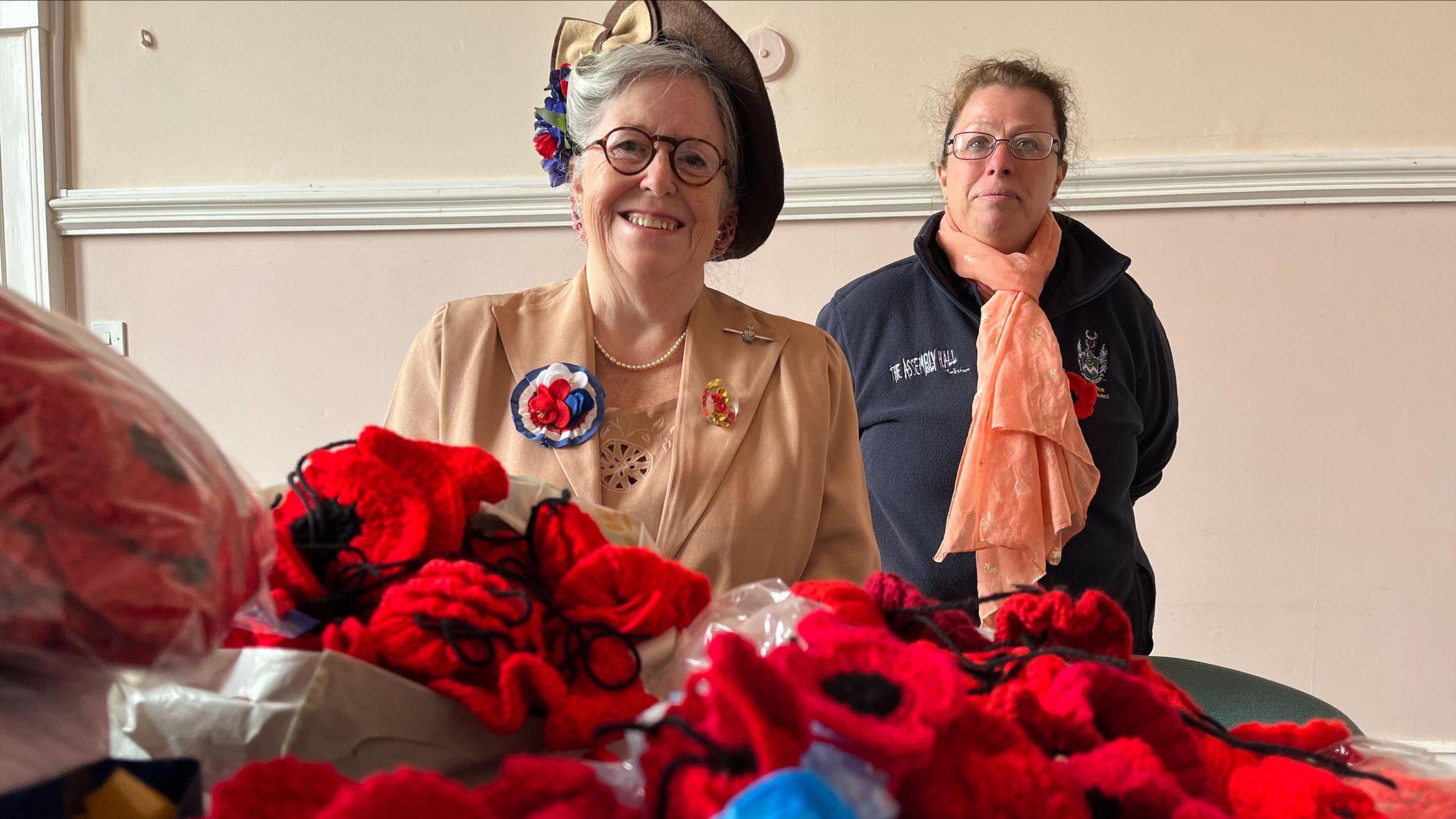 Two women look at the camera, one smiling, one placid, with knitted poppies lying about in the foreground.