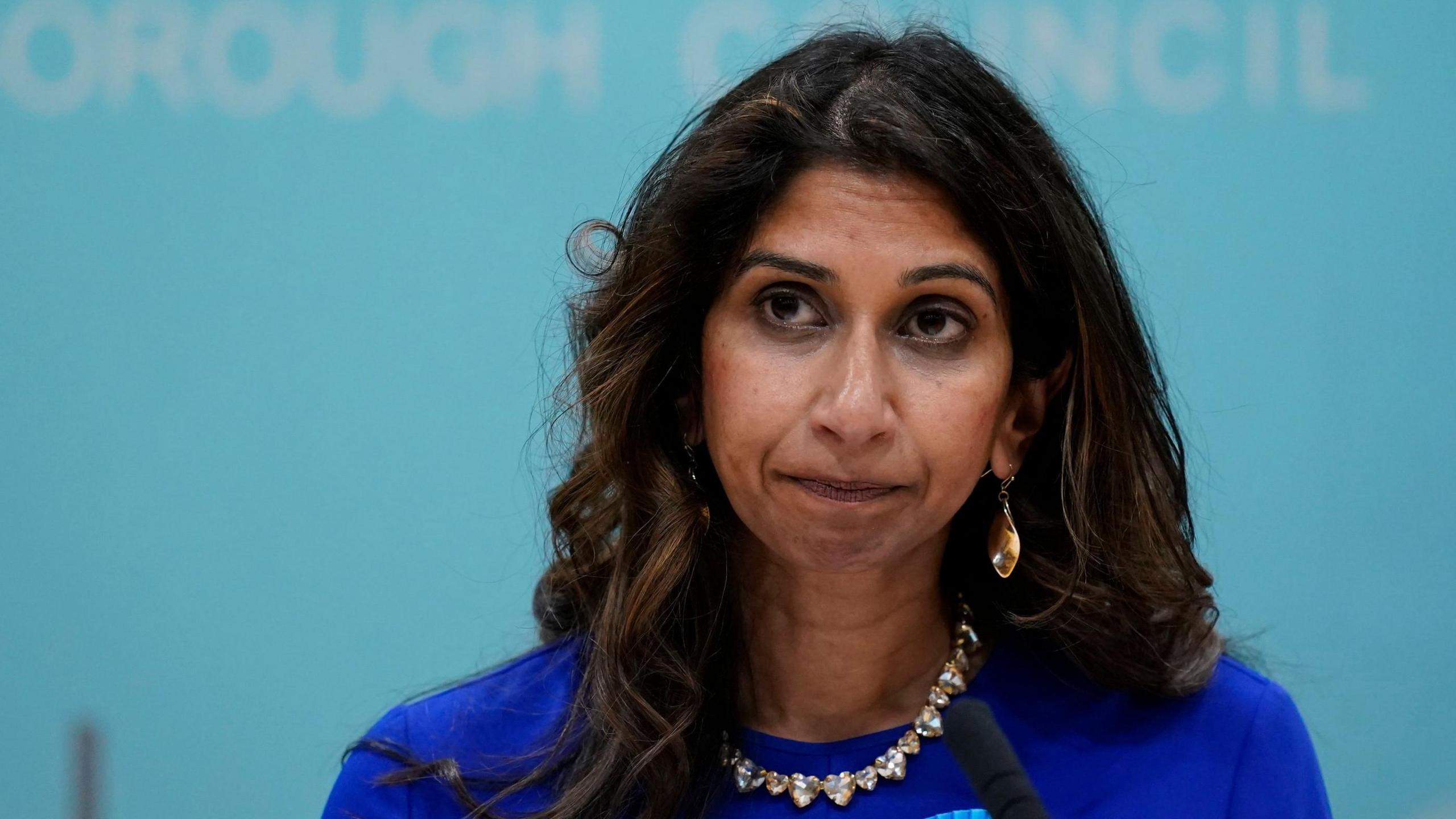 Suella Braverman MP is pictured with a serious expression in a council room. She has wavy, mid-length, dark hair and wears a blue top, jewelled necklace and earrings.