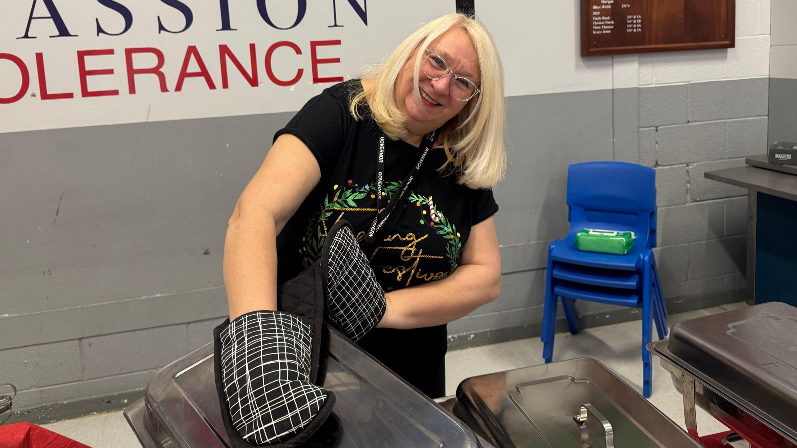 Angela, a woman with long blonde hair, standing in the kitchen smiling with oven gloves, about to remove the lid from a catering tray