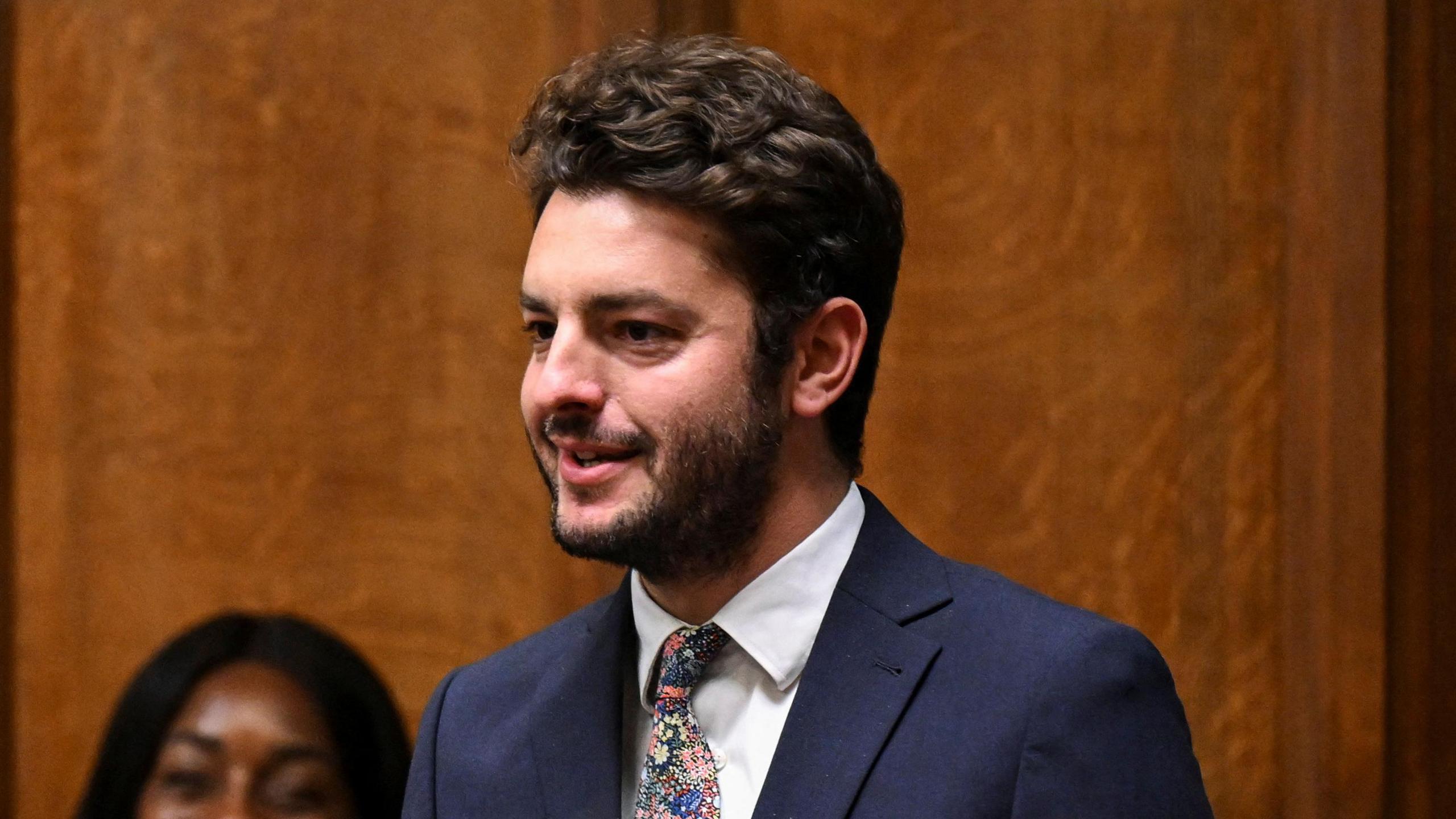 MP Jack Abbott speaking in the House of Commons. He is wearing a navy suit jacket, white shirt and has a patterned tie. He has dark hair and a beard.