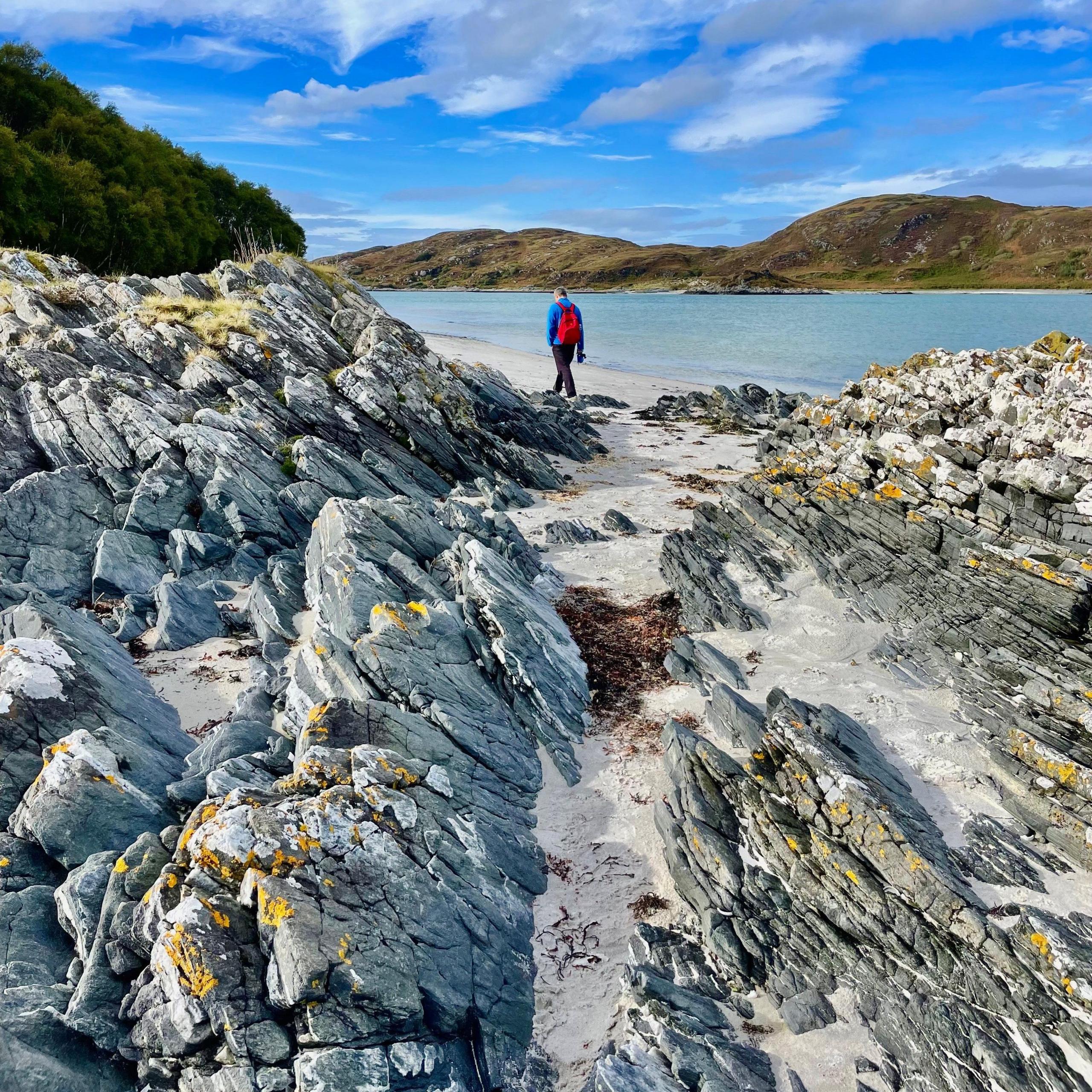 A rocky outcrop is in the foreground. In the background is the sea and hills. The sky is bright blue