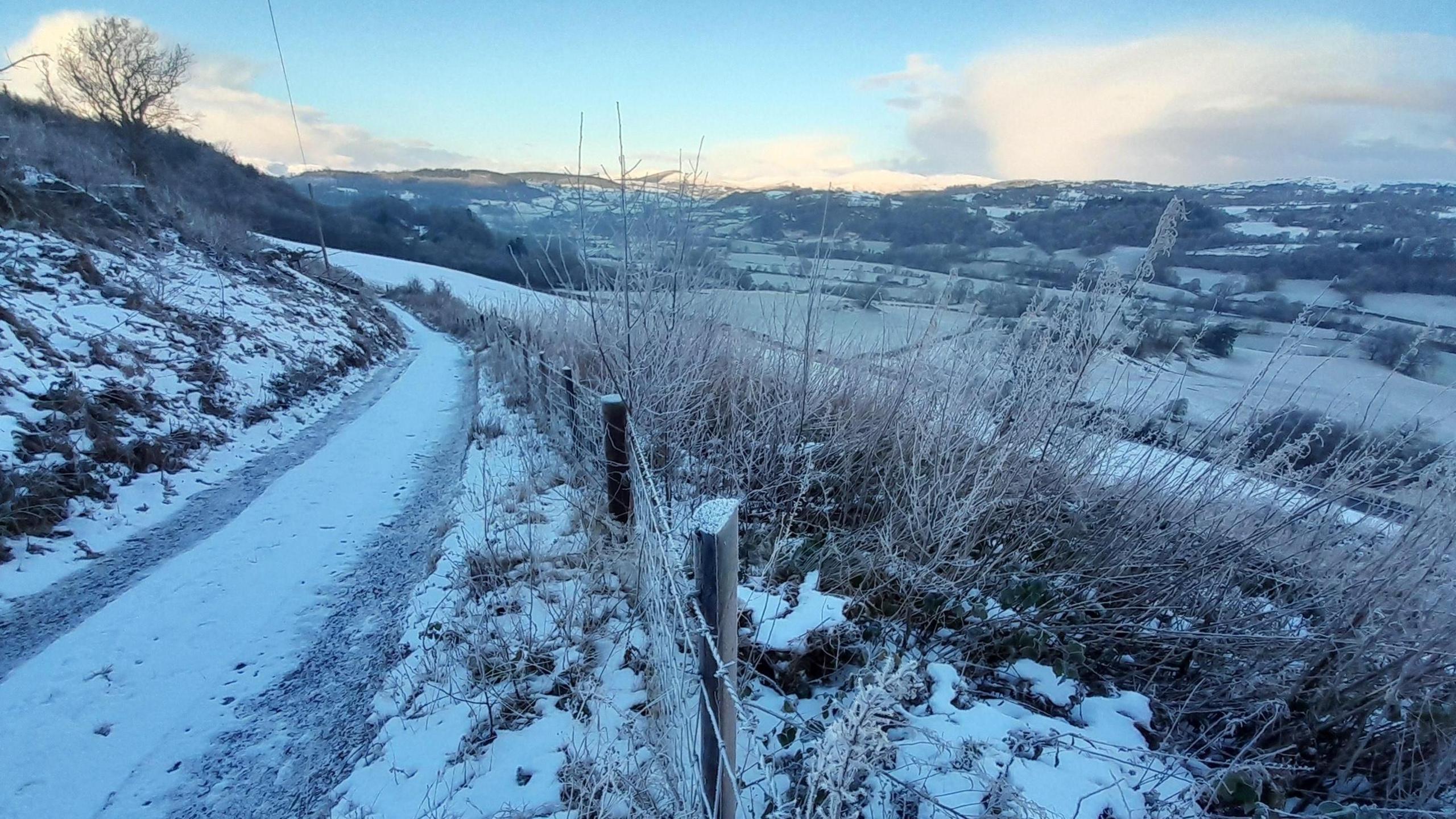 Rural road covered in snow with vehicle tracks and field sloping down to the right. Snow-covered hills are in the background.