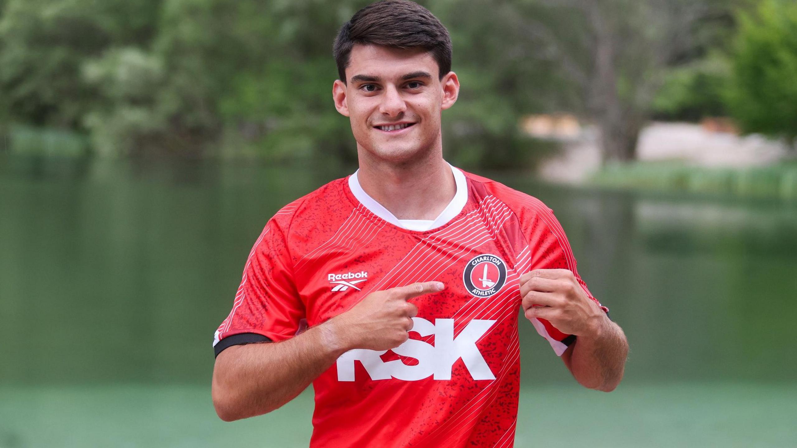 Rob Apter wearing a red Charlton Athletic kit, pointing at the club crest with his right hand and holding his shirt with his left hand, standing in front of a lake