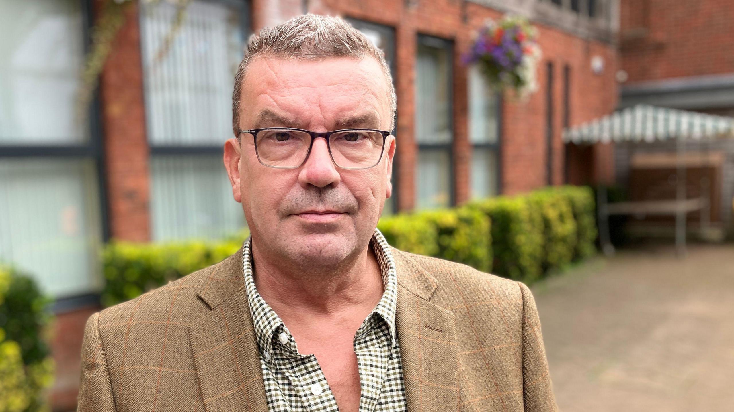 A short grey haired man, wearing a brown blazer and check shirt, standing in front of a red-bricked building, with tall windows and cropped green bushes.