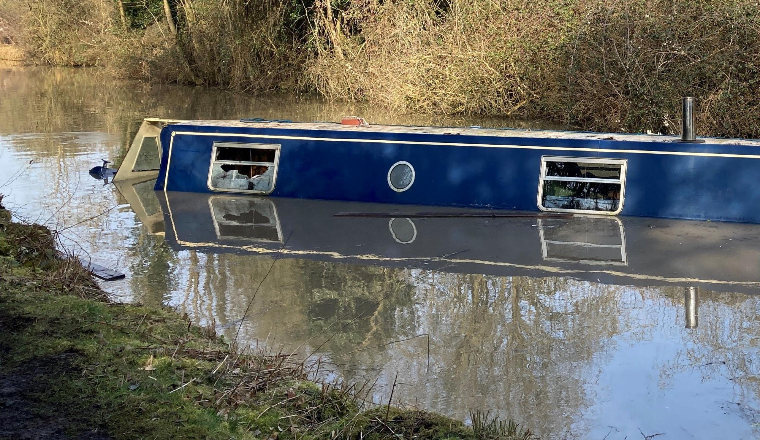 A semi-submerged blue narrowboat sits in brown canal water. A window has been smashed. There is greenery on either side of the banks.