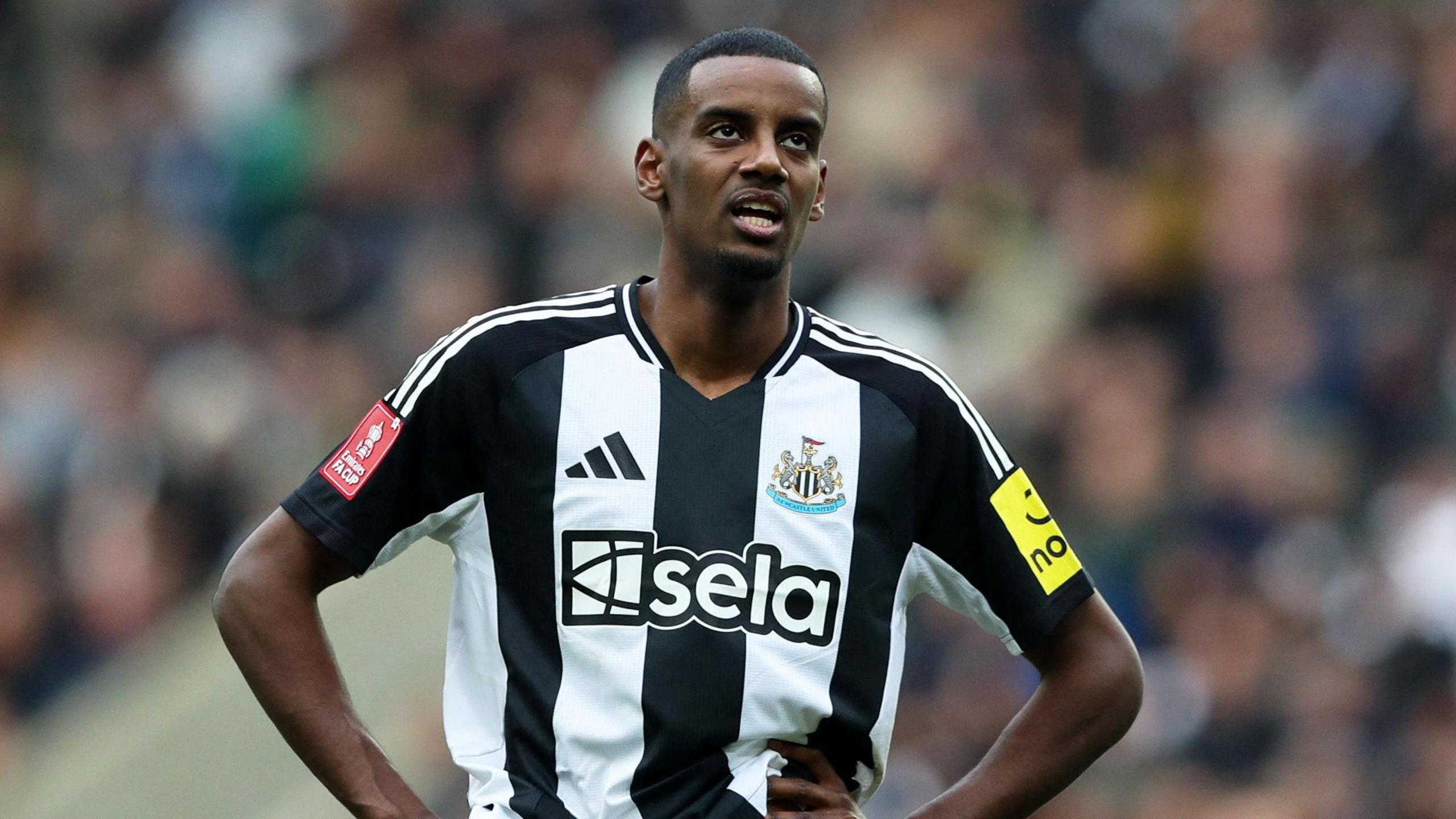 Isak playing football. He is standing with his hands on his waist and is looking skyward. He is wearing a black and white Newcastle United shirt.