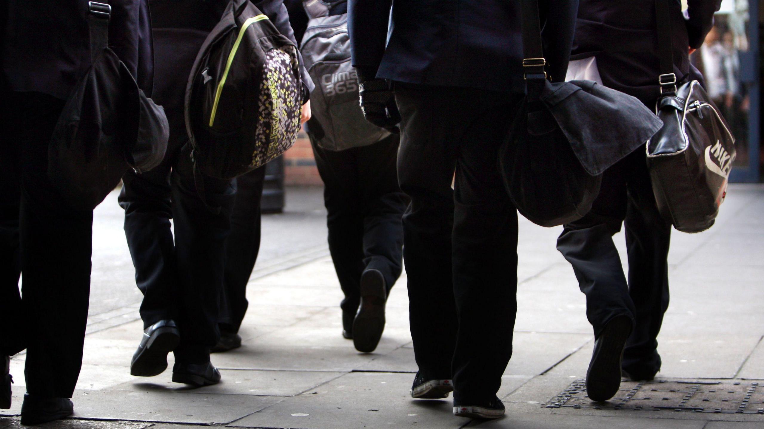 School children walking into school. The image shows the legs of pupils carrying large school bags.