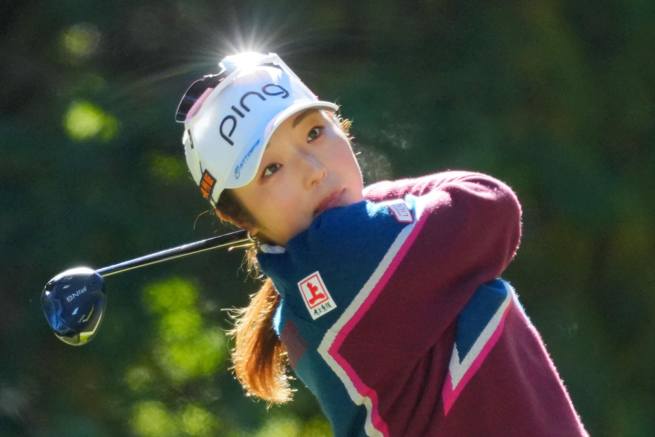 A golfer follows through on a powerful swing, wearing a white cap and maroon-blue sweater, with sunlight glinting off her visor against a soft, blurred green background.