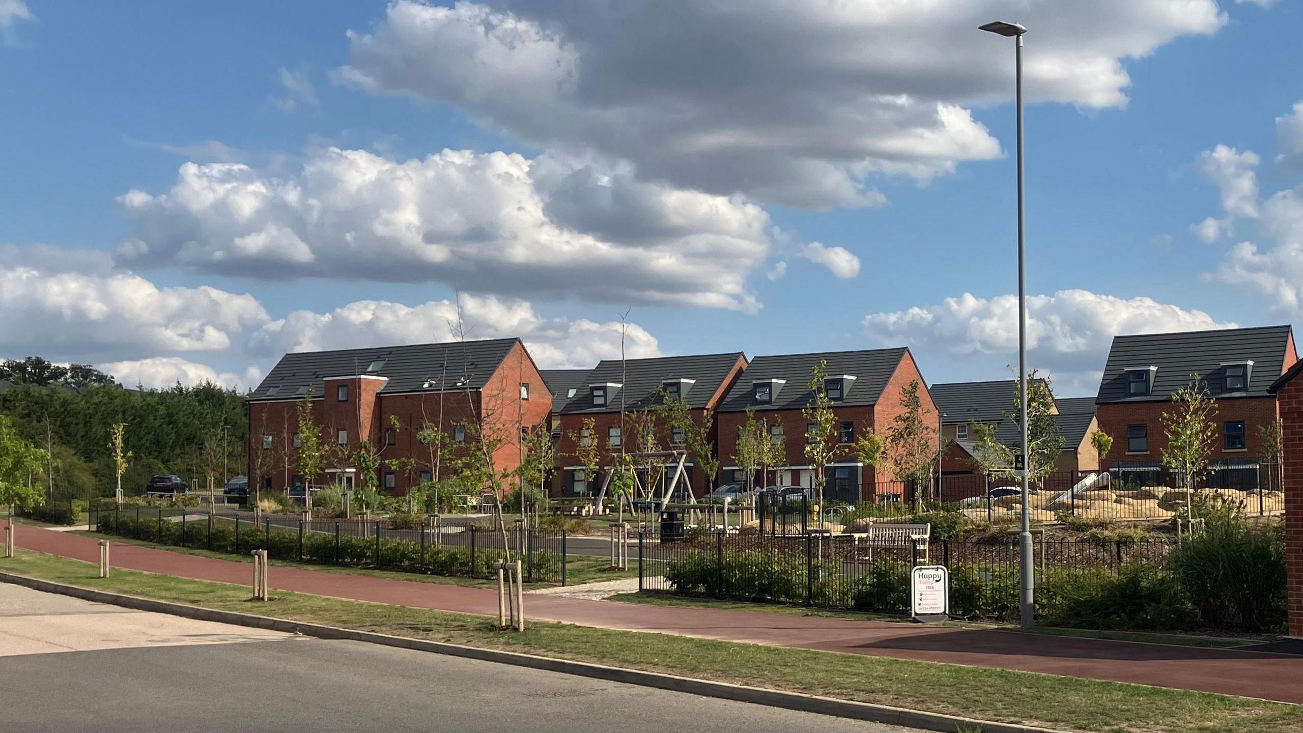 A housing estate with red brick homes in the background. In front of them is a children's play area where swings can be seen. In front of that a footpath, a grass verge which has small trees on it, and then a road. A large part of the picture is a light blue sky with large fluffy white clouds.