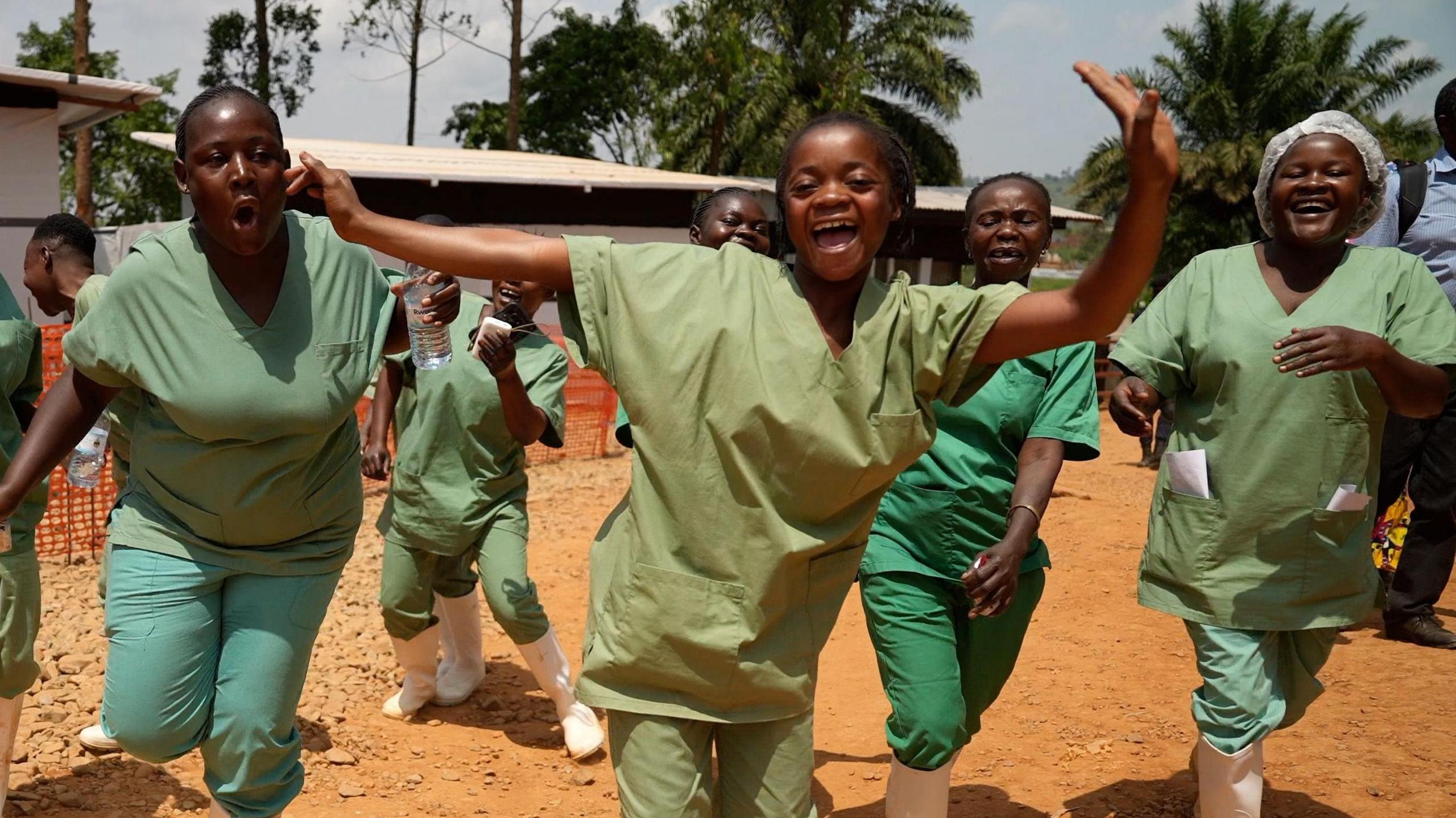 Health workers wearing green scrubs and white wellie boots dance and sing on a dusty ground
