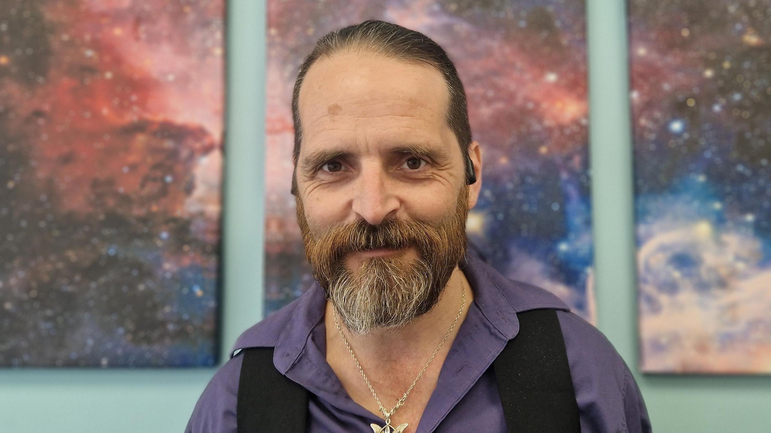 Rob has dark hair tied back in a pony tail and a beard. He is wearing a purple shirt which is open at the collar, a black waistcoat and a large silver pendant necklace. He is standing in front of three canvasses of the night sky. It is a head and shoulders shot of him. 