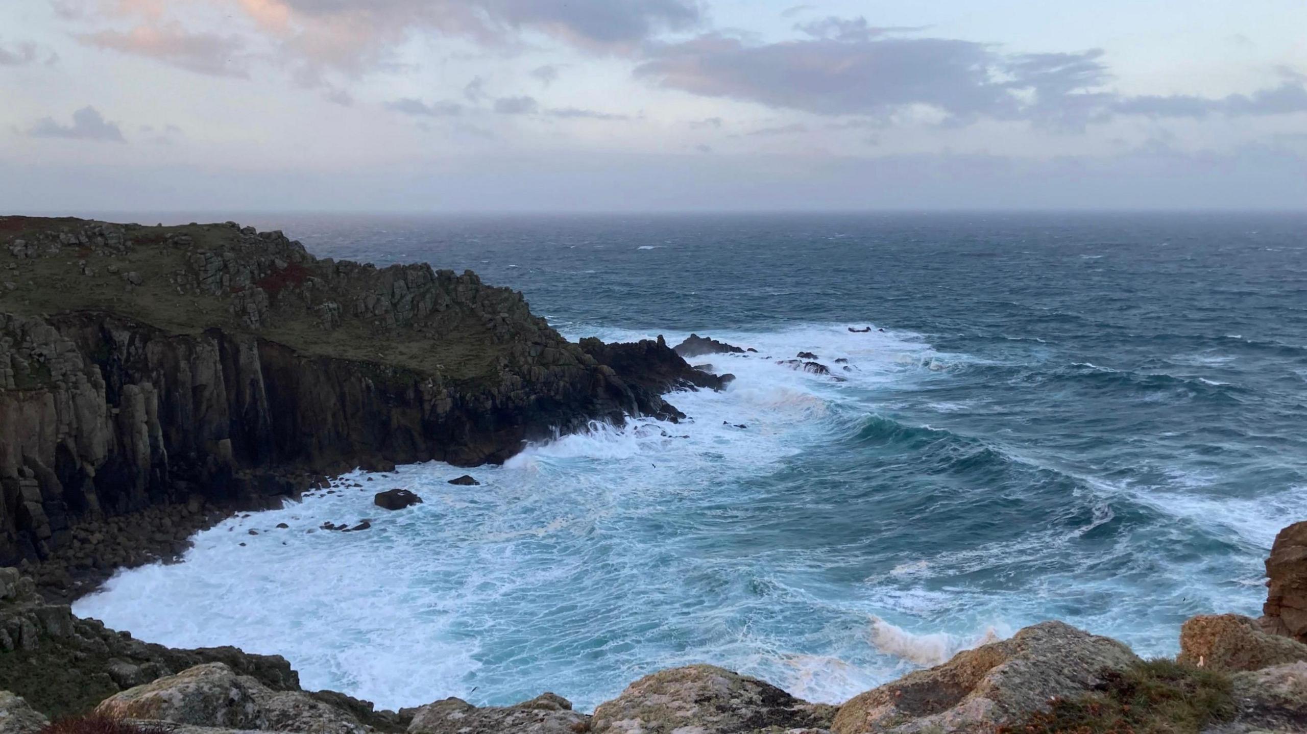 A picture of the sea at St Levan. The U-shaped rugged coastline can be seen next to the body of water which is breaking as it hits the rocks. The sky is a pale blue colour with a number of clouds pictured.