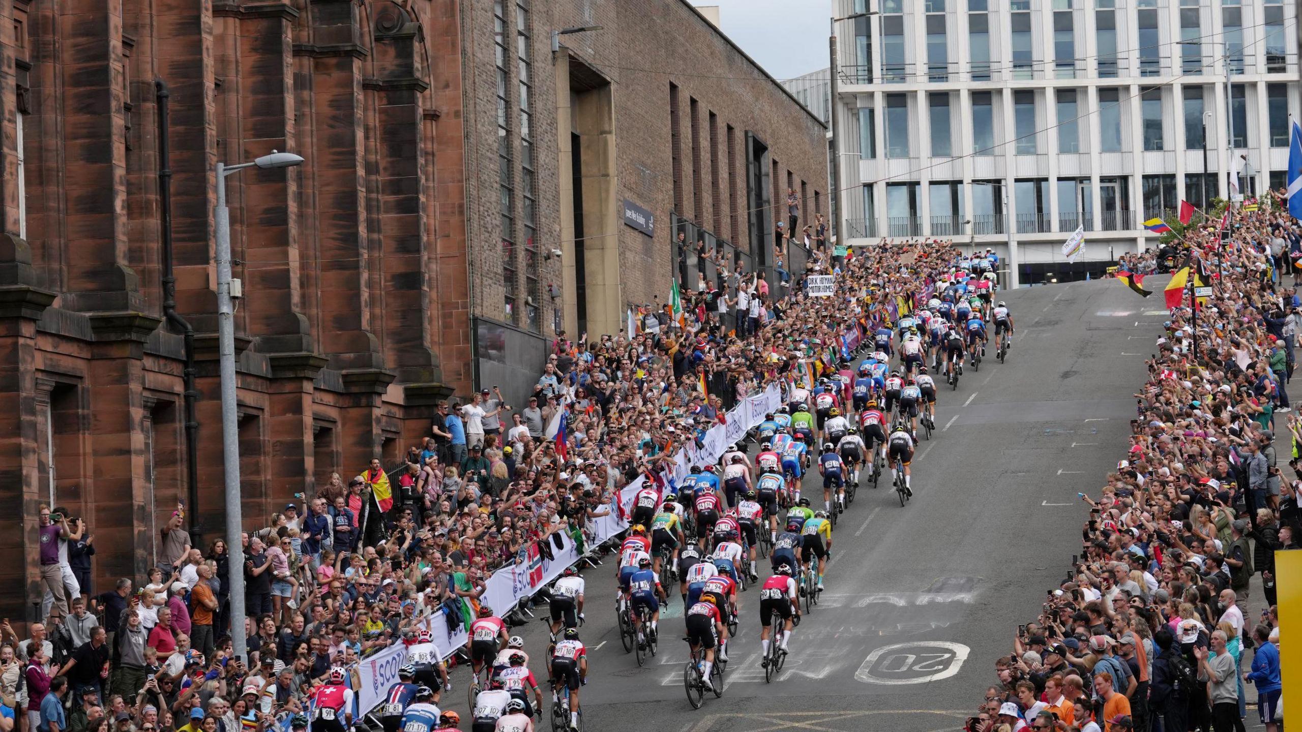 Dozens of cyclists going up Montrose Street during the men's elite road race at the UCI Cycling World Championships. The pavements are packed with people cheering them on, waving flags